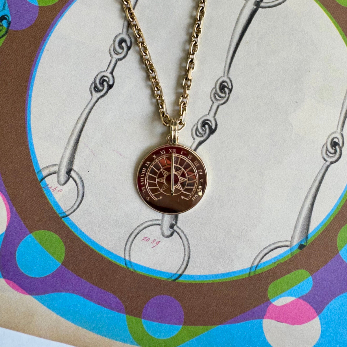 A gold chain necklace with a round pendant featuring a red and gold compass design, displayed on a colorful vintage-style illustrated card.