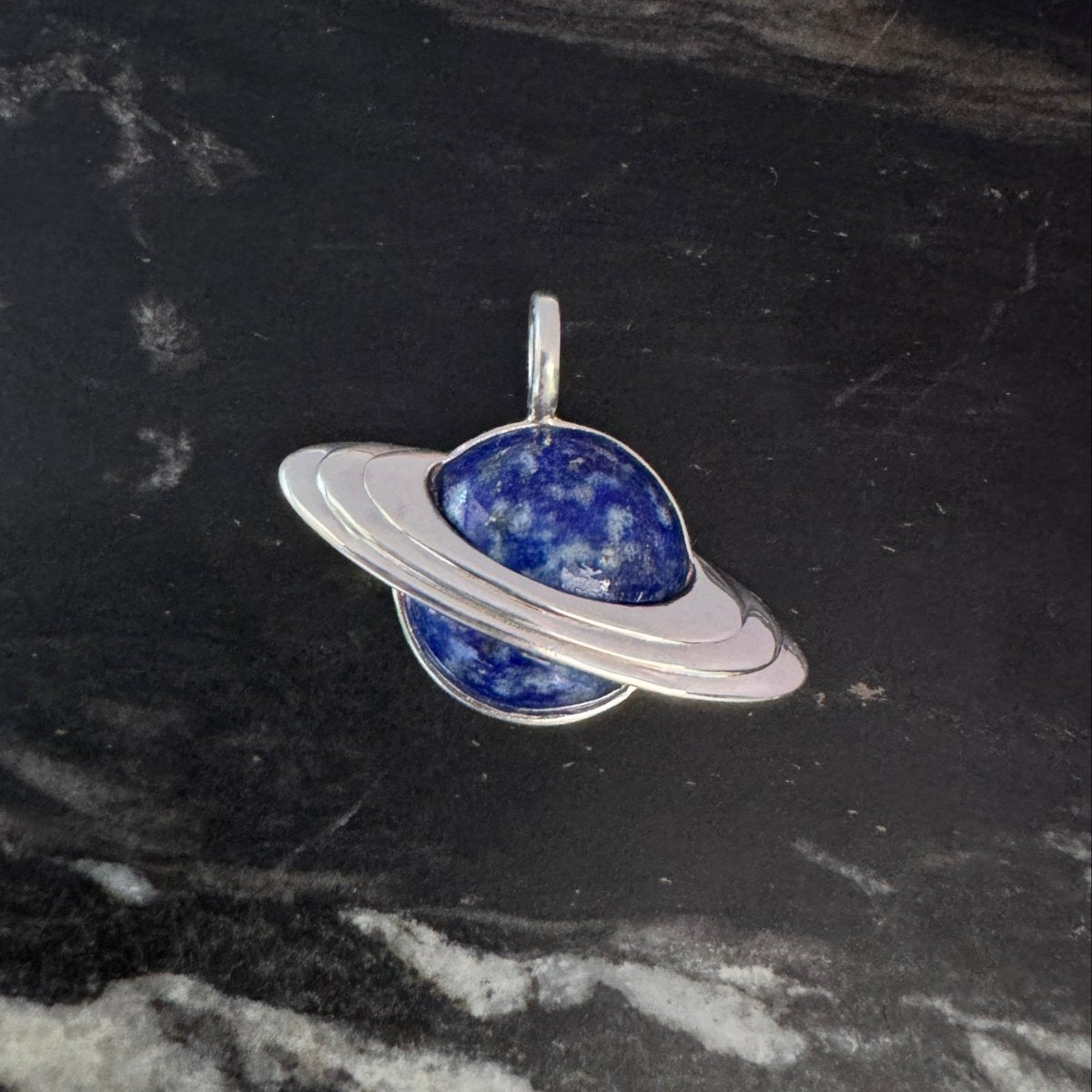 Silver Saturn-shaped pendant with blue lapis inlay against a black and white marble background