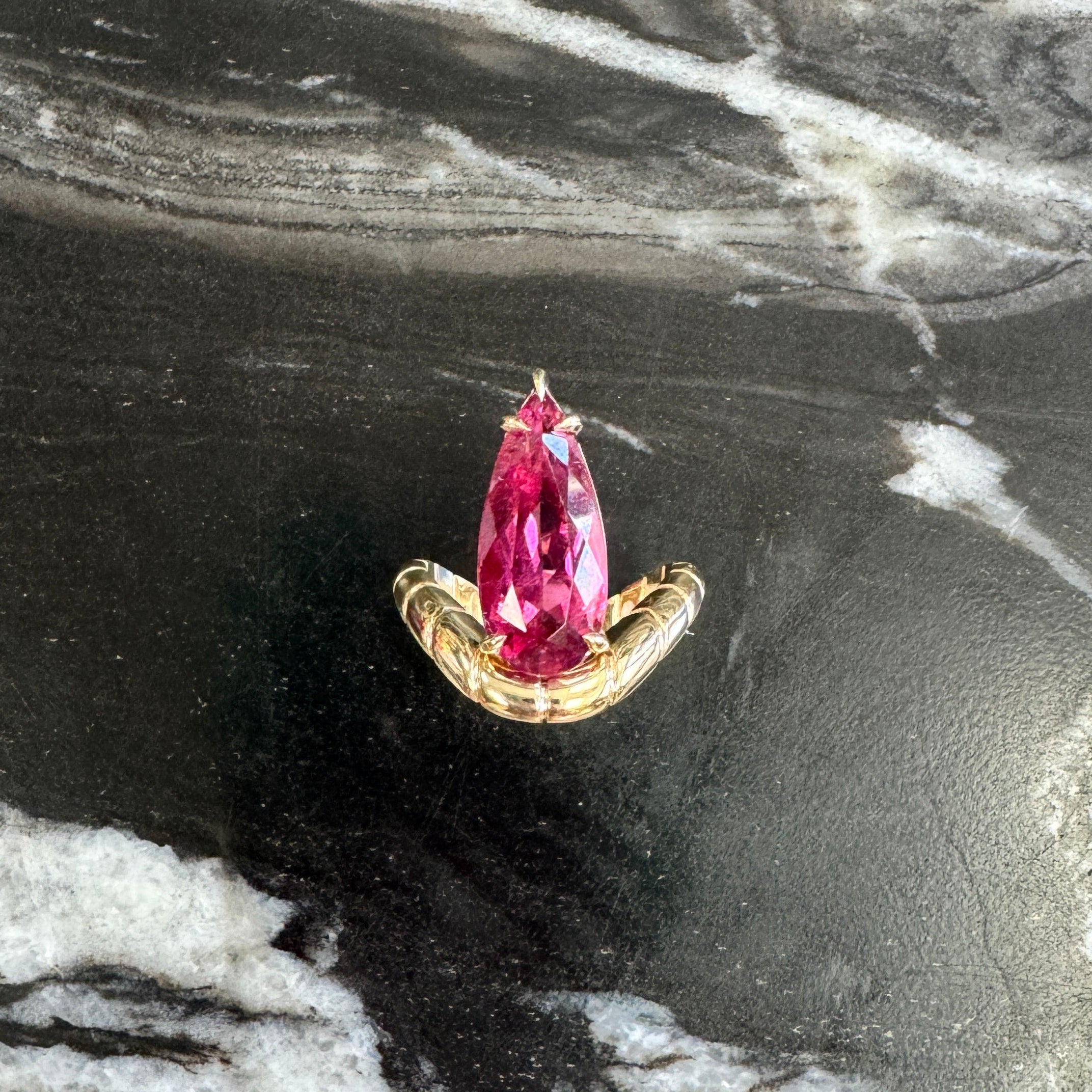Gold ring with a curved band and a pear shaped pink rubellite against a black and white marble background 