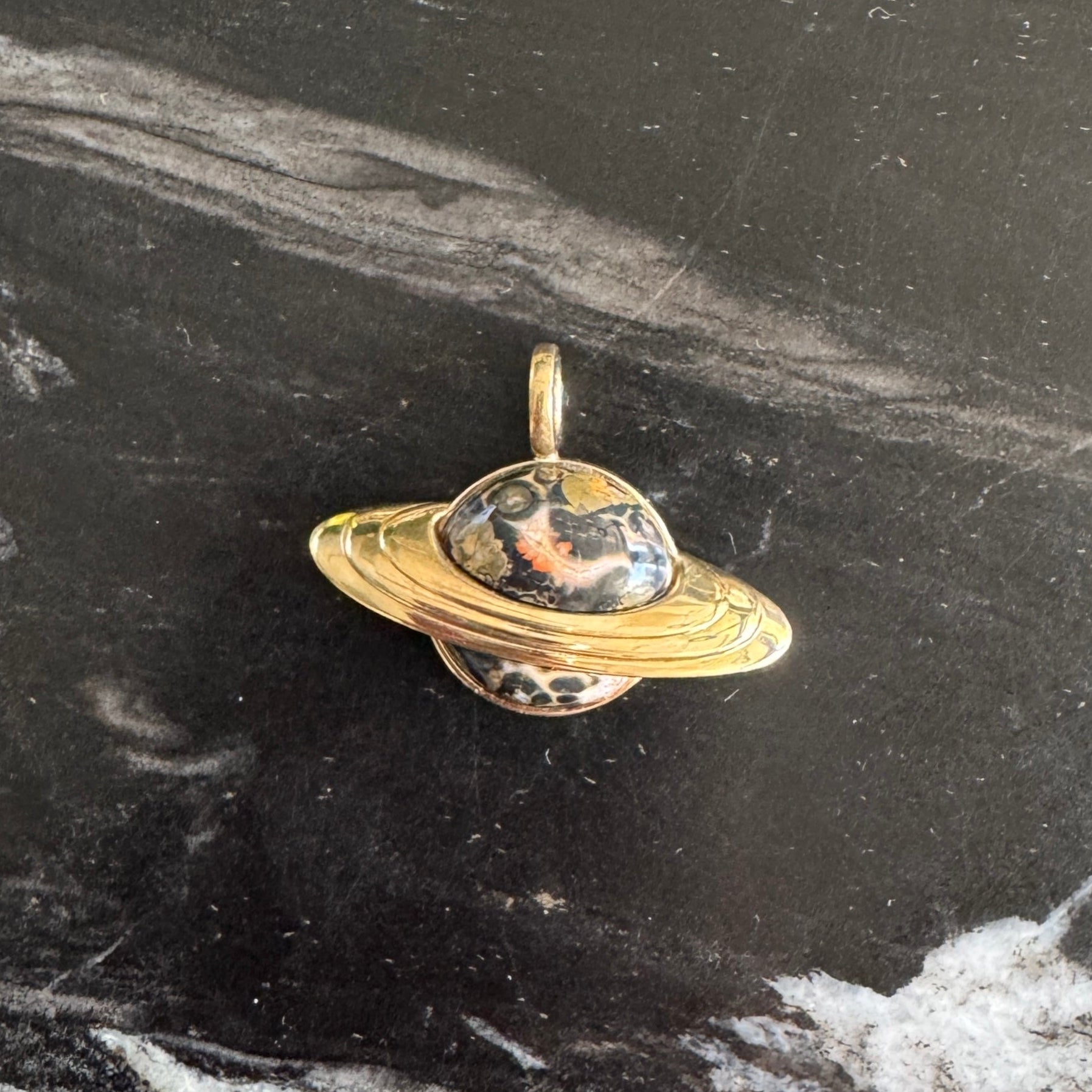 Gold Saturn-shaped pendant with leopard Jasper inlay against a black and white marble background 