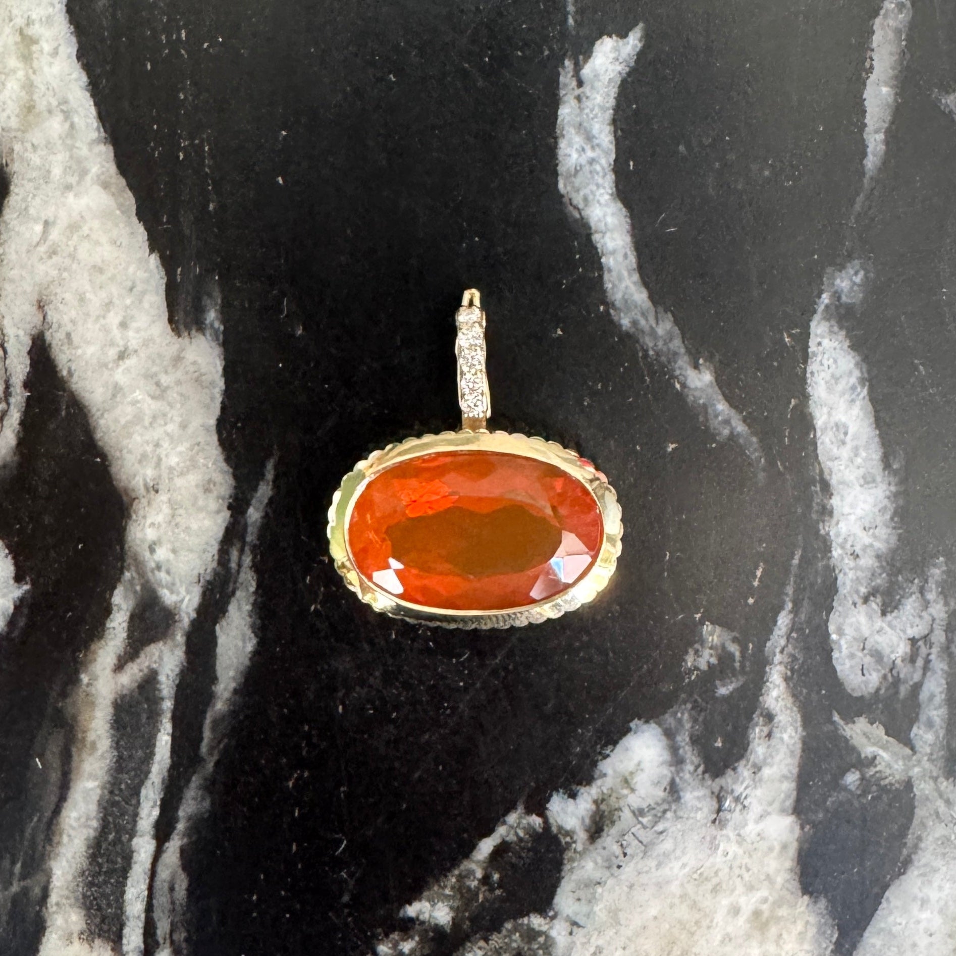 Gold pendant with an orange oval fire opal and a diamond bail on a black and white marble surface 
