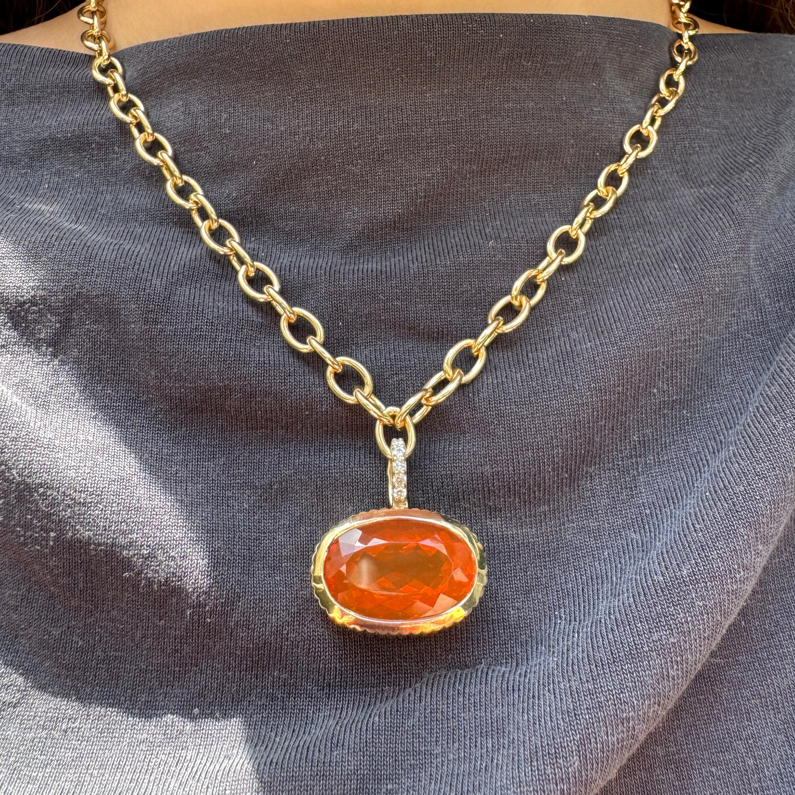 Person wearing a gold pendant with an orange oval fire opal and a diamond bail on a gold chain against a gray top