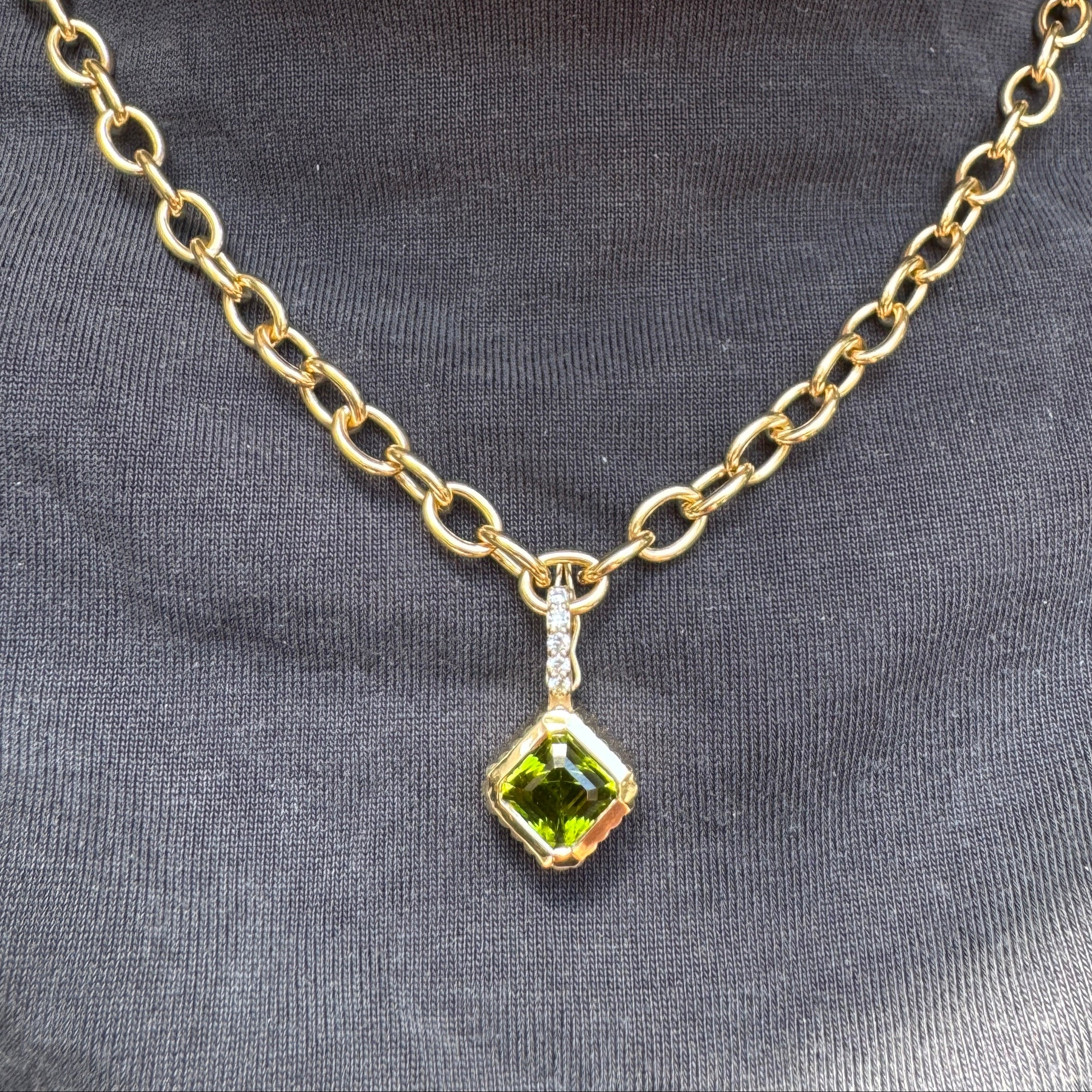 Person wearing a gold pendant with a square cut green peridot and a diamond bail on a gold chain against gray shirt