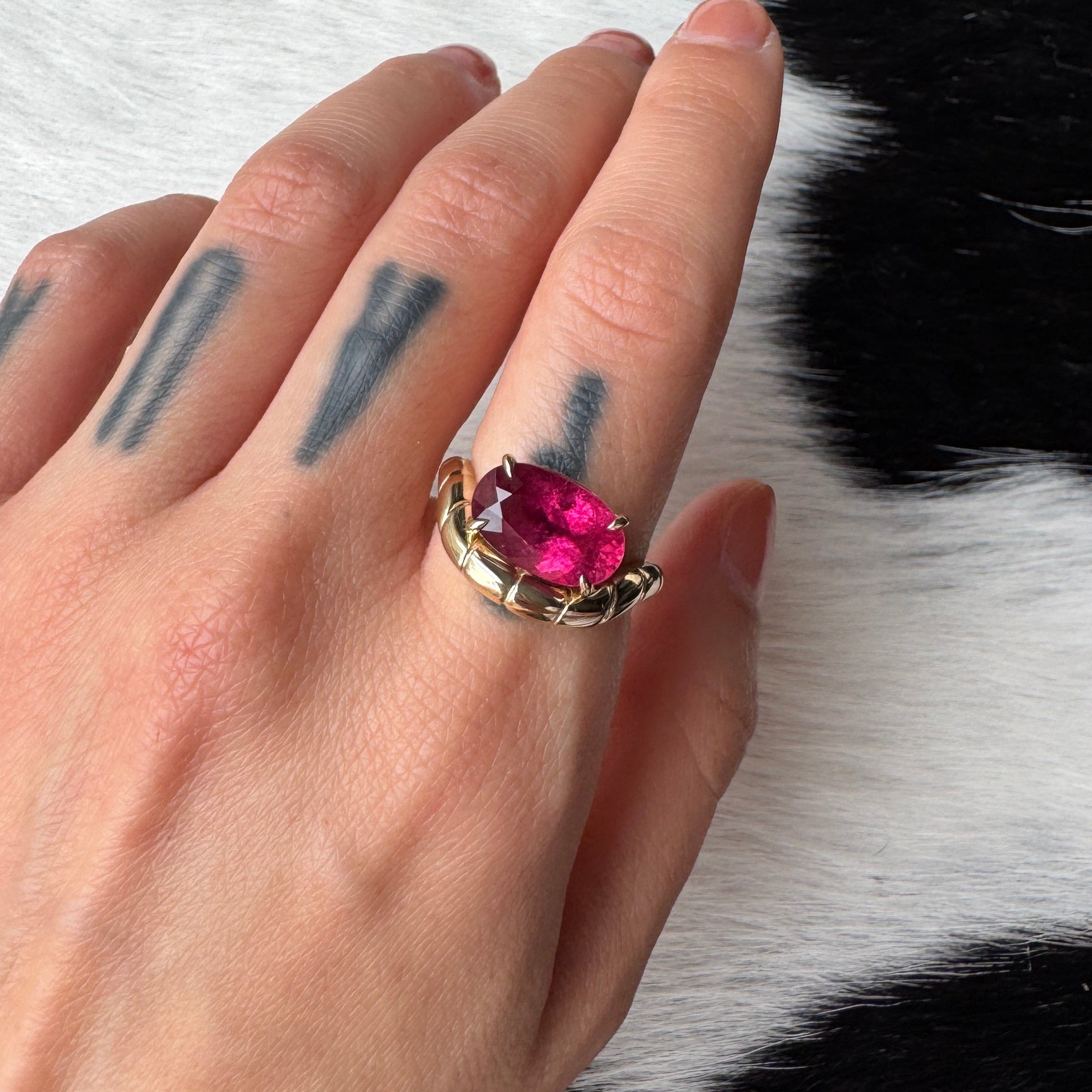Hand wearing a gold ring with a curved band and an oval pink rubellite against a black and white cow print background 