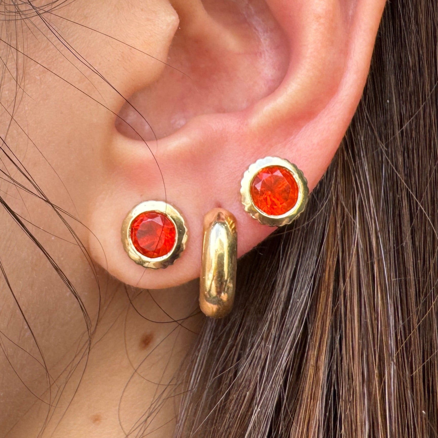 Close-up of an ear wearing gold earrings including two round gold studs with orange fire opals