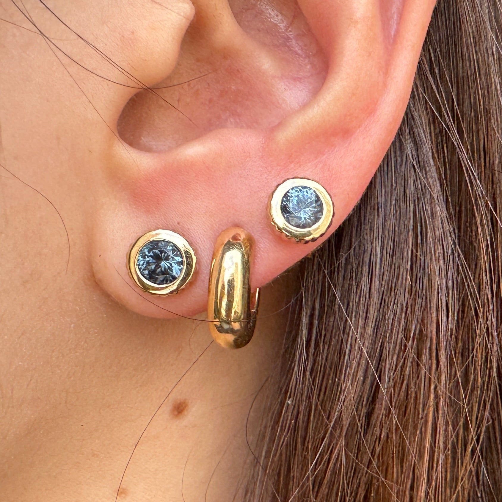 Close-up of an ear wearing gold earrings including two round gold studs with blue spinels 