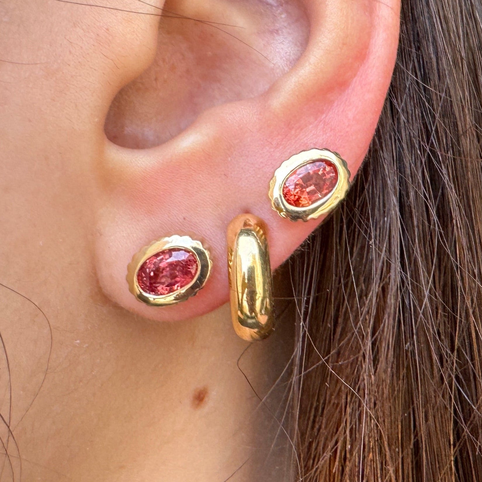 Close-up of an ear wearing gold earrings including two gold oval studs with pinky orange sapphires