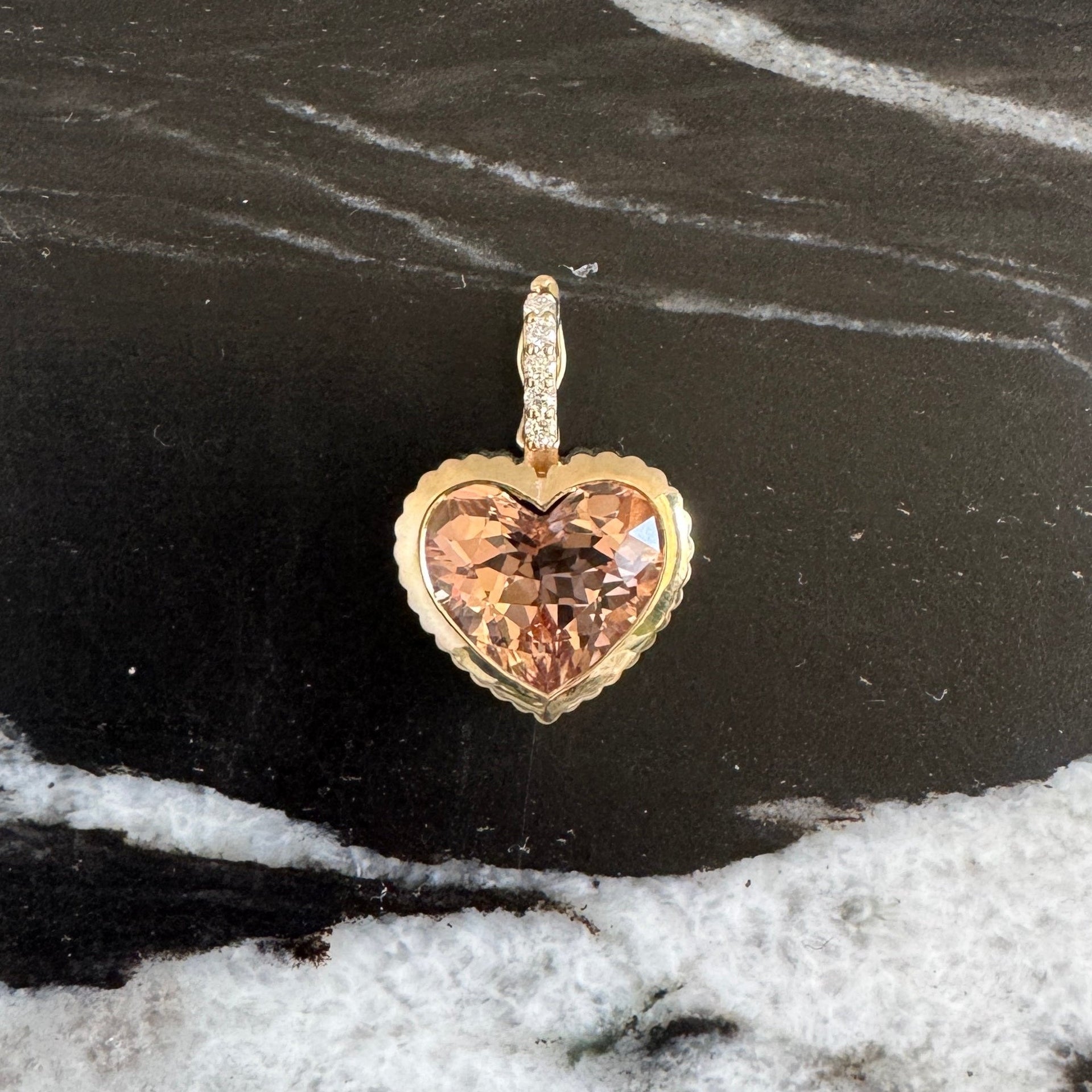 Gold heart-shaped pendant with a peachy morganite and a diamond bail against a black and white marble background 