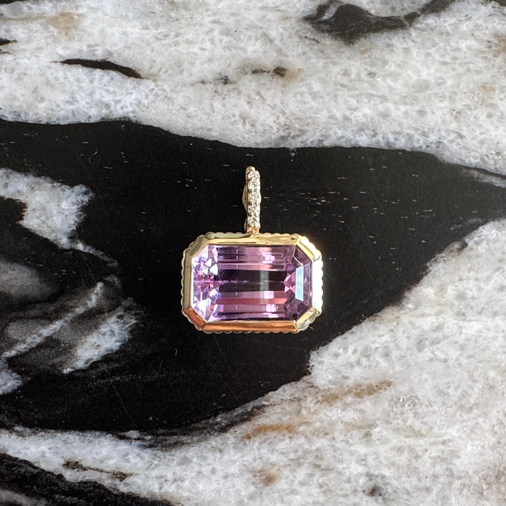 Gold pendant with a purple kunzite  and a diamond bail against a black and white  marble background 