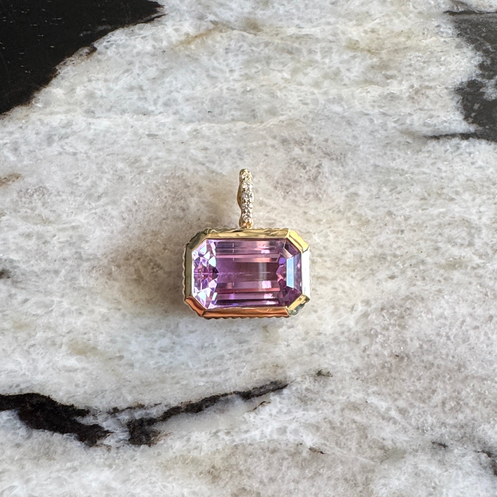 Gold pendant with a purple kunzite  and a diamond bail against a black and white  marble background 