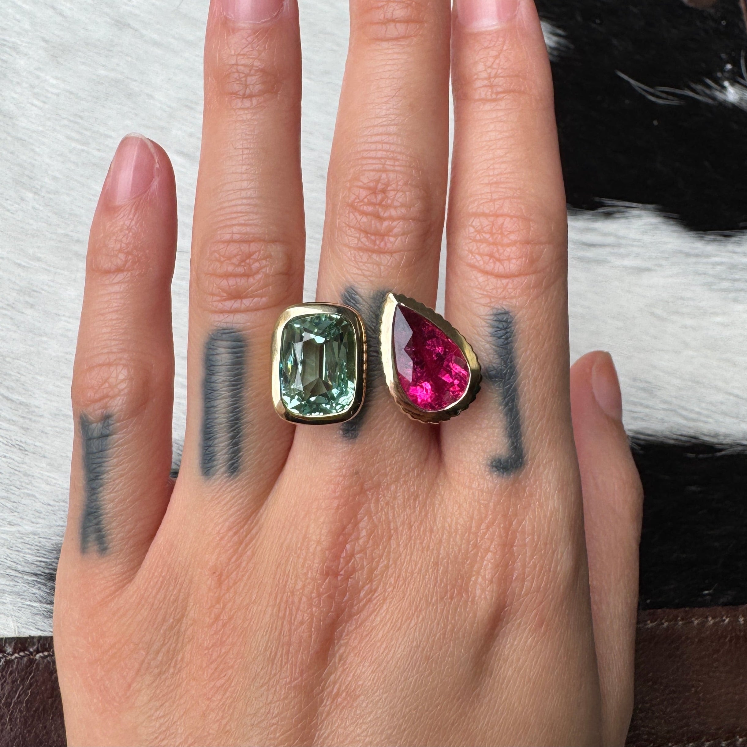 Hand wearing a gold ring with an elongated cushion-cut mint tourmaline and a pear-shaped pink red rubellite against a black and white cow print background