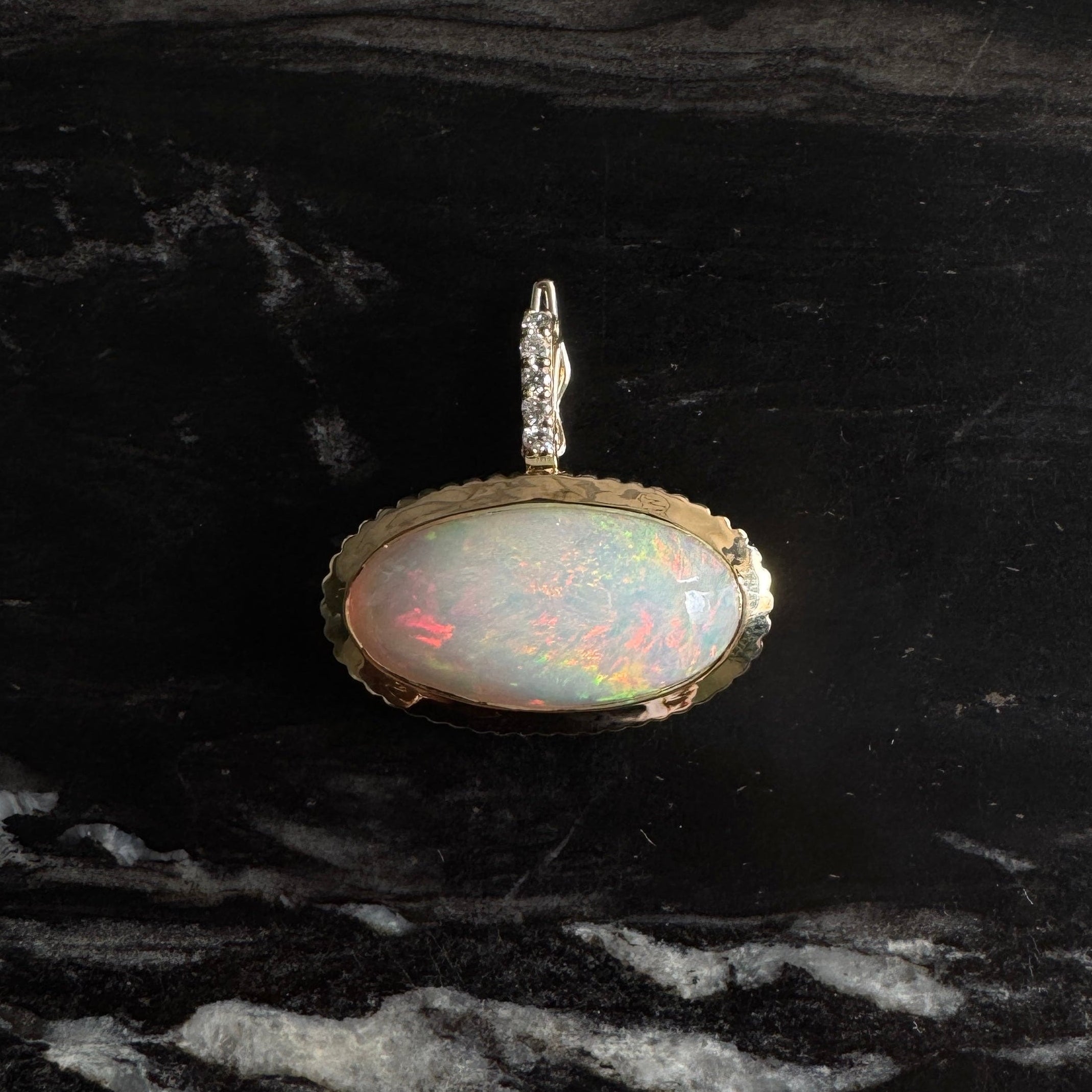 Gold pendant with a rainbow cabochon opal and a diamond bail against a black and white marble background