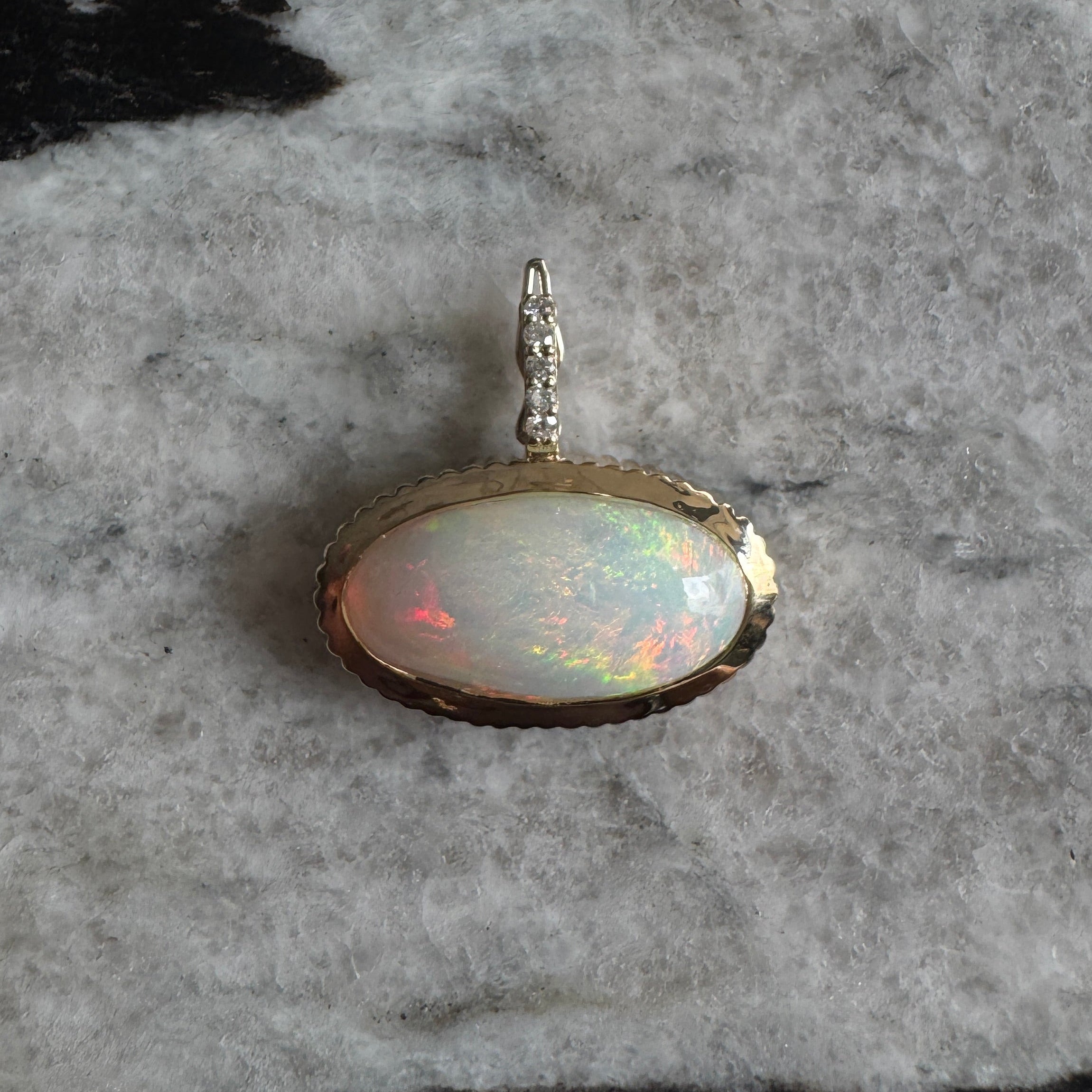 Gold pendant with a rainbow cabochon opal and a diamond bail against a black and white marble background