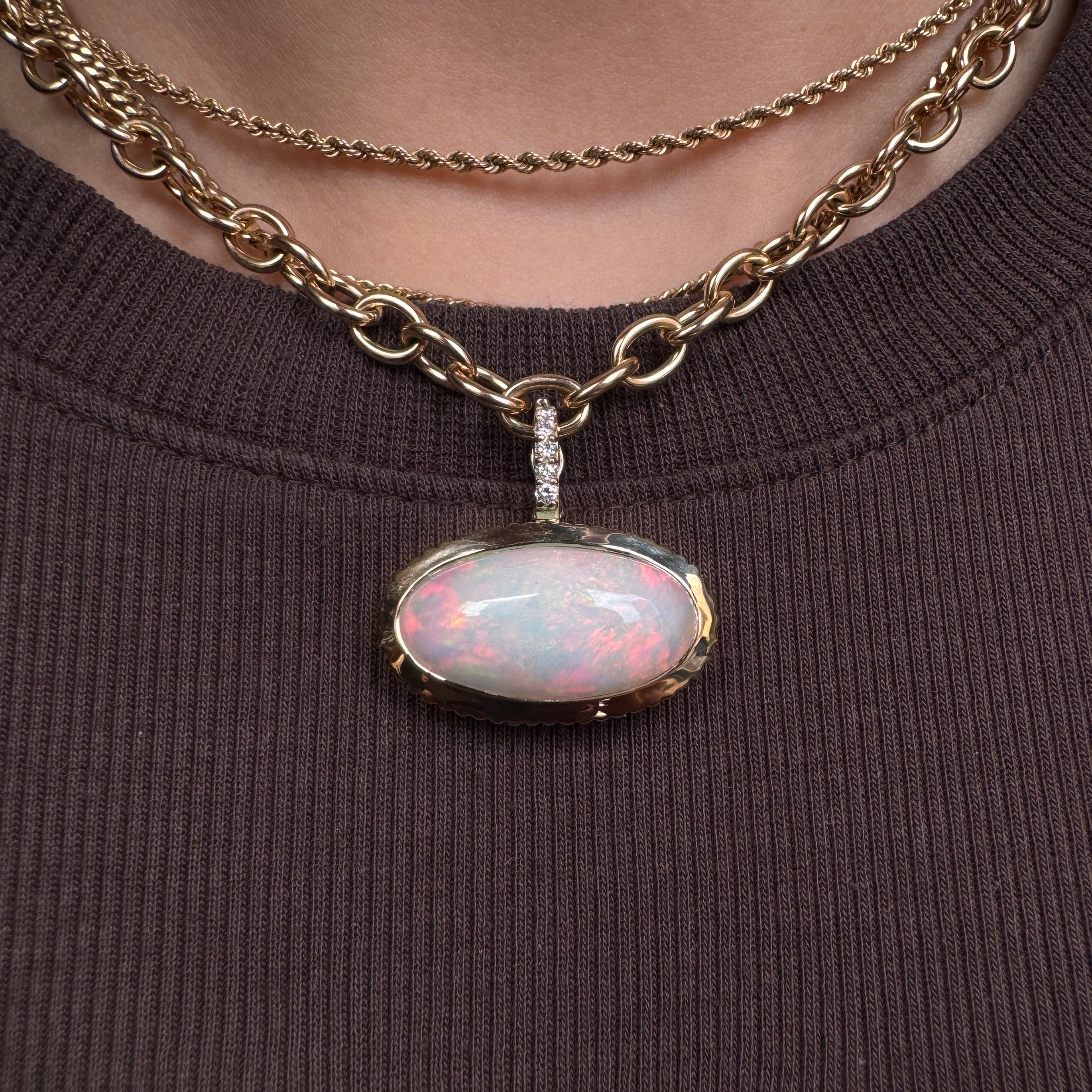 Person wearing a gold pendant with a rainbow cabochon opal and a diamond bail against a brown top
