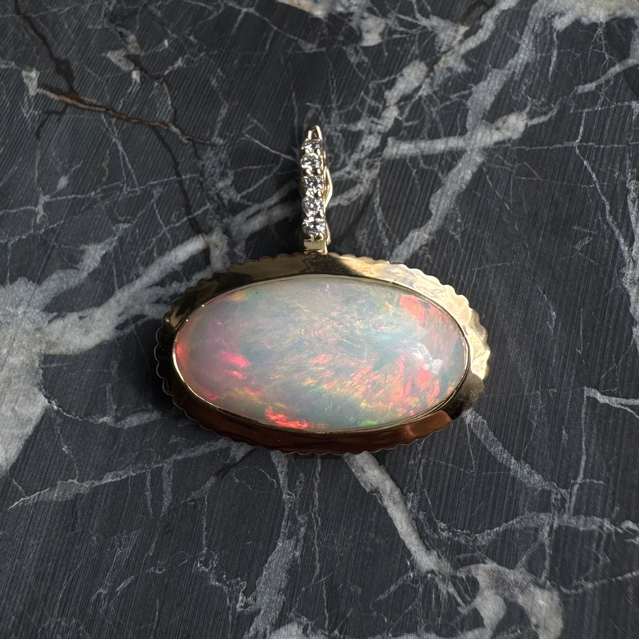 Gold pendant with a rainbow cabochon opal and a diamond bail against a black and white marble background