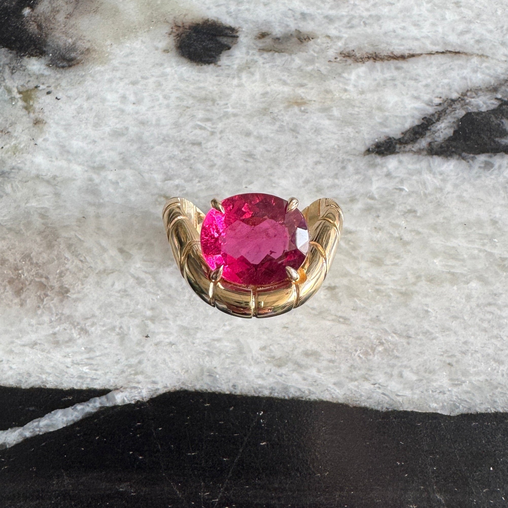 Gold ring with oval-cut rubellite and a curved band against a black and white marble background 
