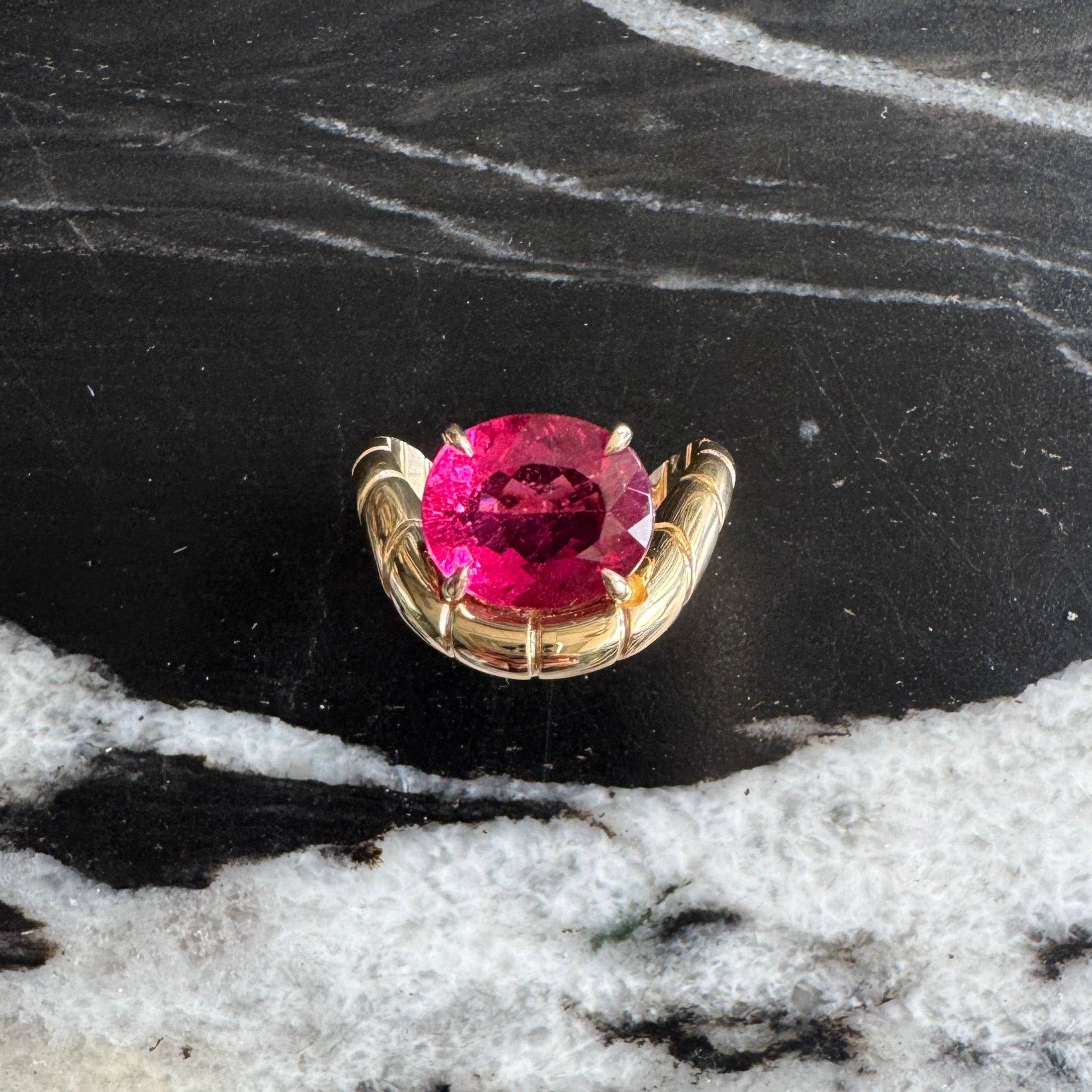 Gold ring with oval-cut rubellite and a curved band against a black and white marble background 