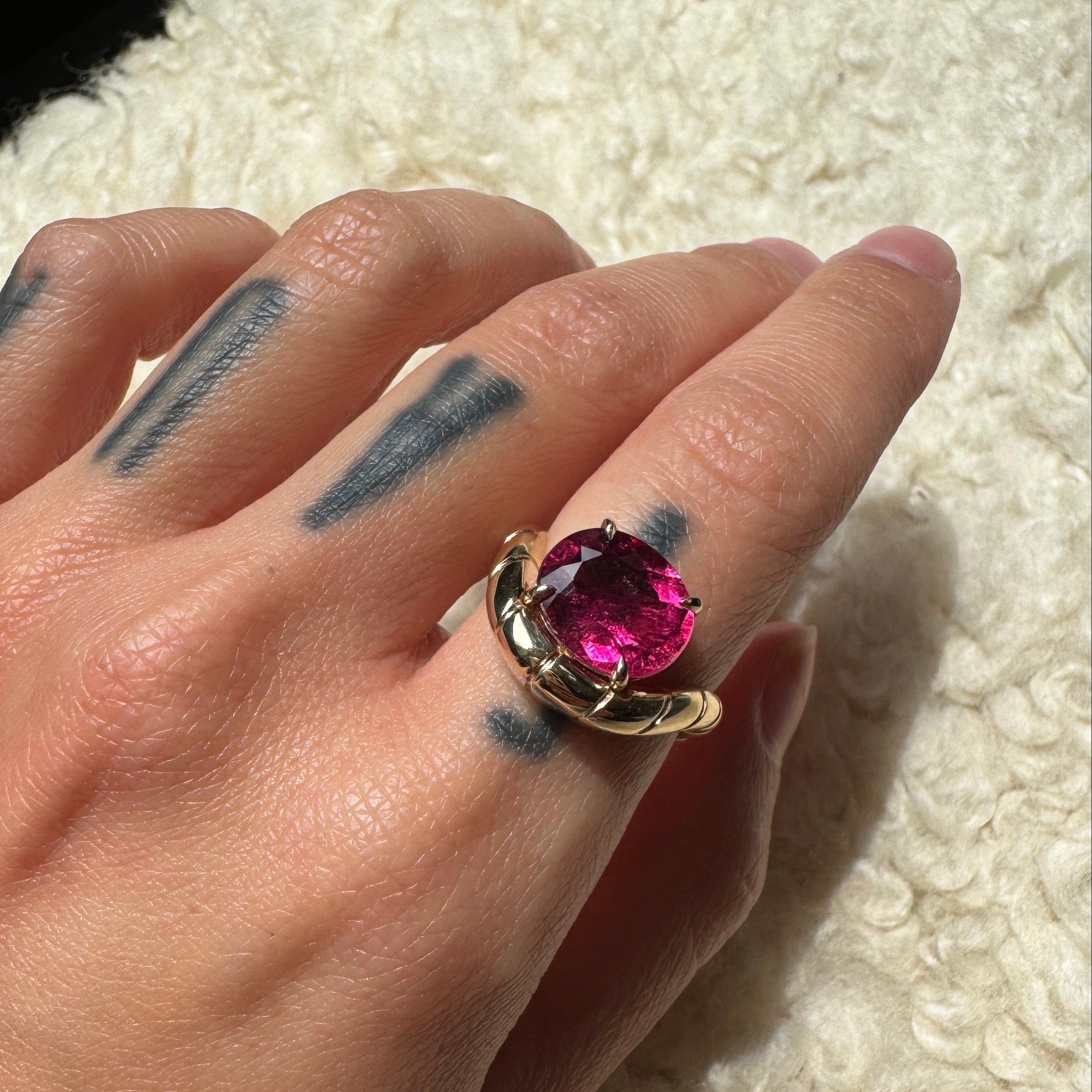 Hand wearing a gold ring with oval-cut rubellite and a curved band against a cream background