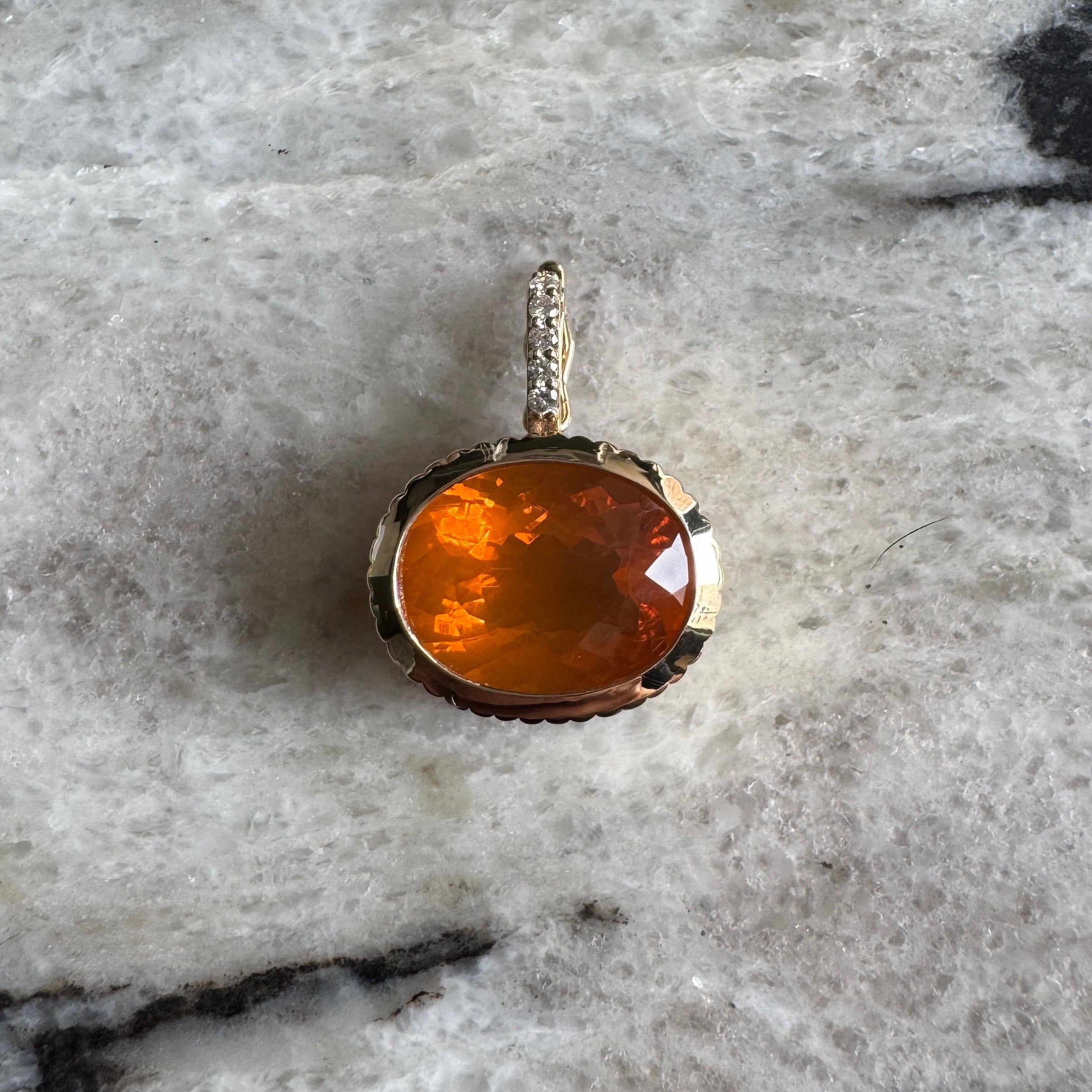 Gold pendant with an oval-cut fire opal and diamond bail against a black and white marble background