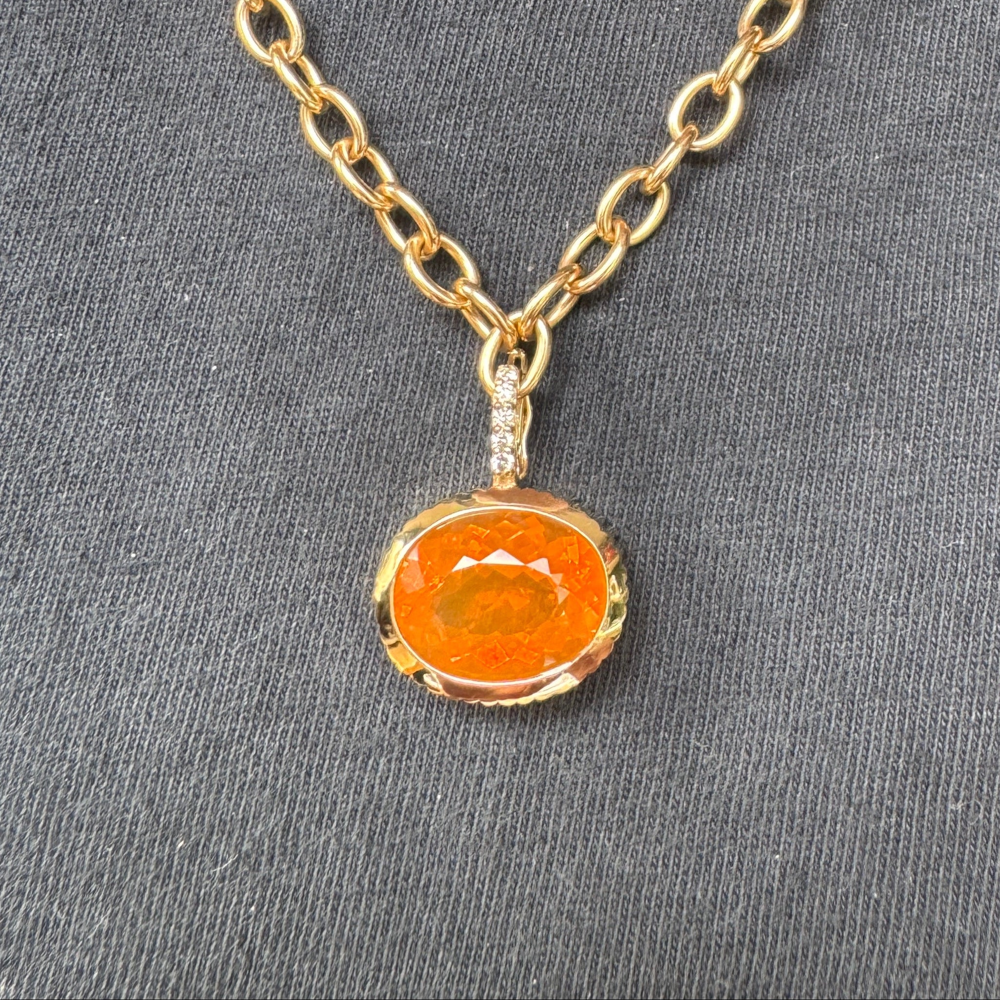 Person wearing a gold pendant with an oval-cut fire opal and diamond bail on a gold chain against a black top