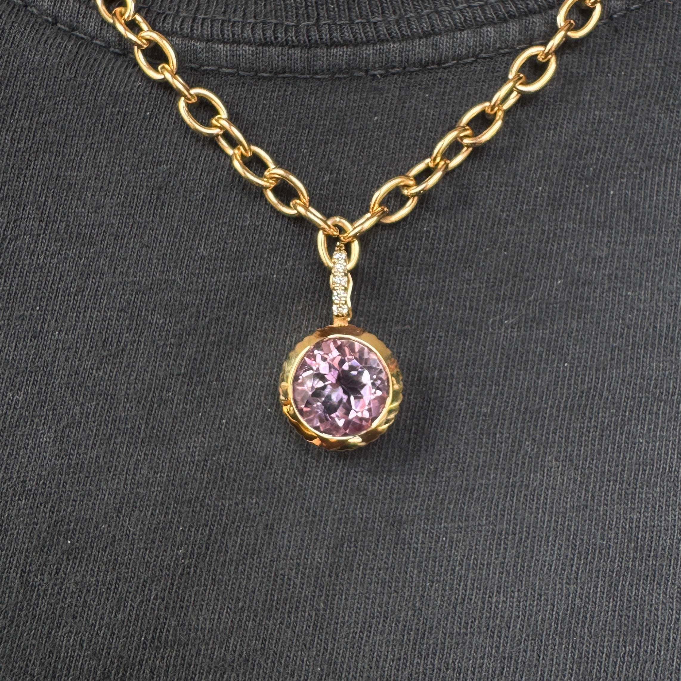 Person wearing a gold pendant with a round purple amethyst and a diamond bail on a gold chain against a black top