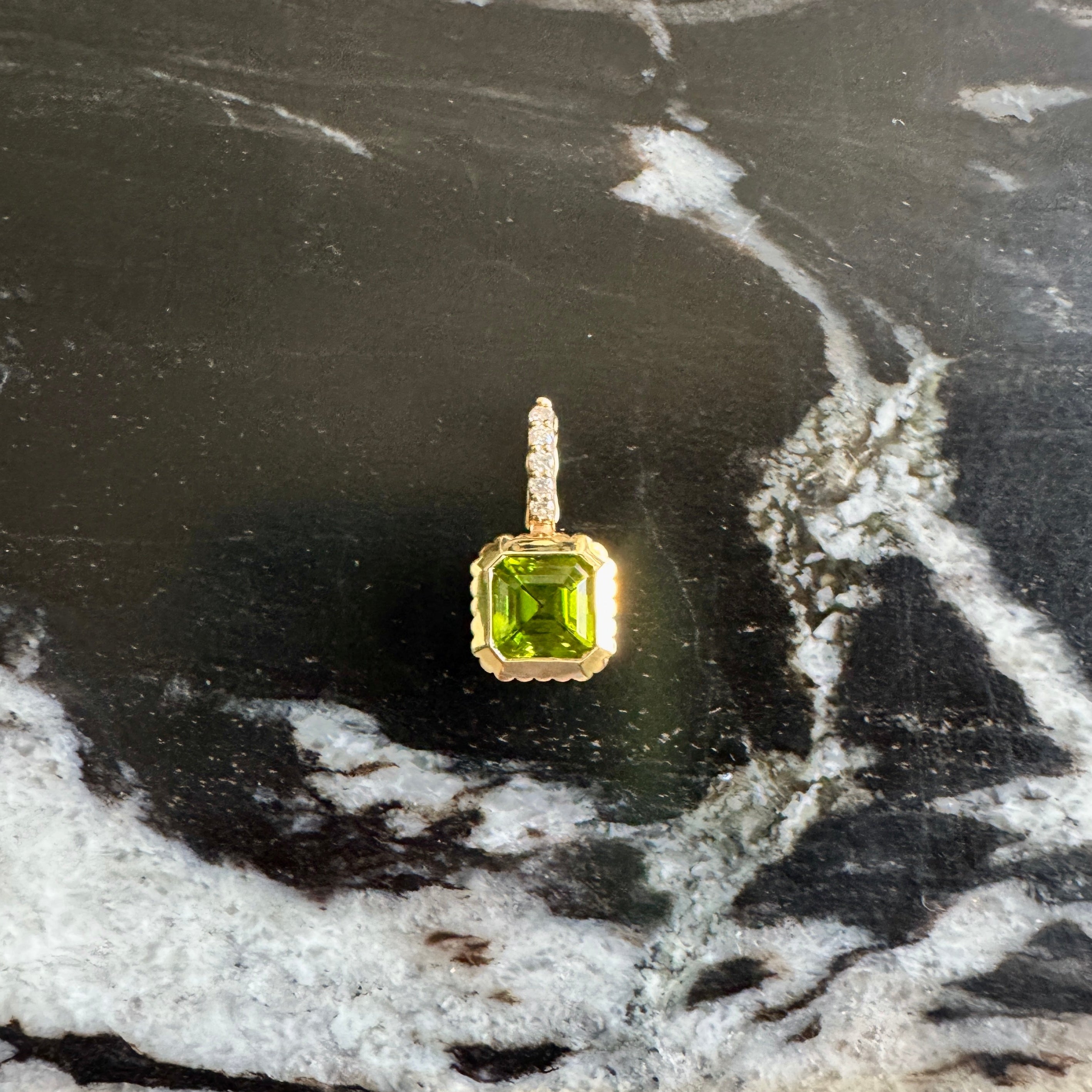 A gold pendant with a square-cut green gemstone, displayed on a black and white marbled surface.