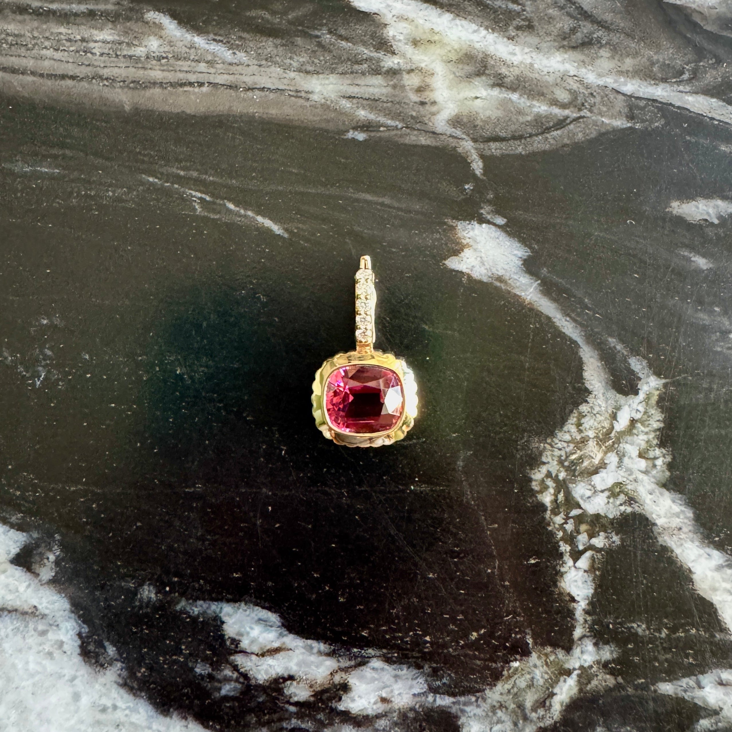 A gold pendant with a cushion-cut red gemstone, displayed on a dark, veined marble surface.