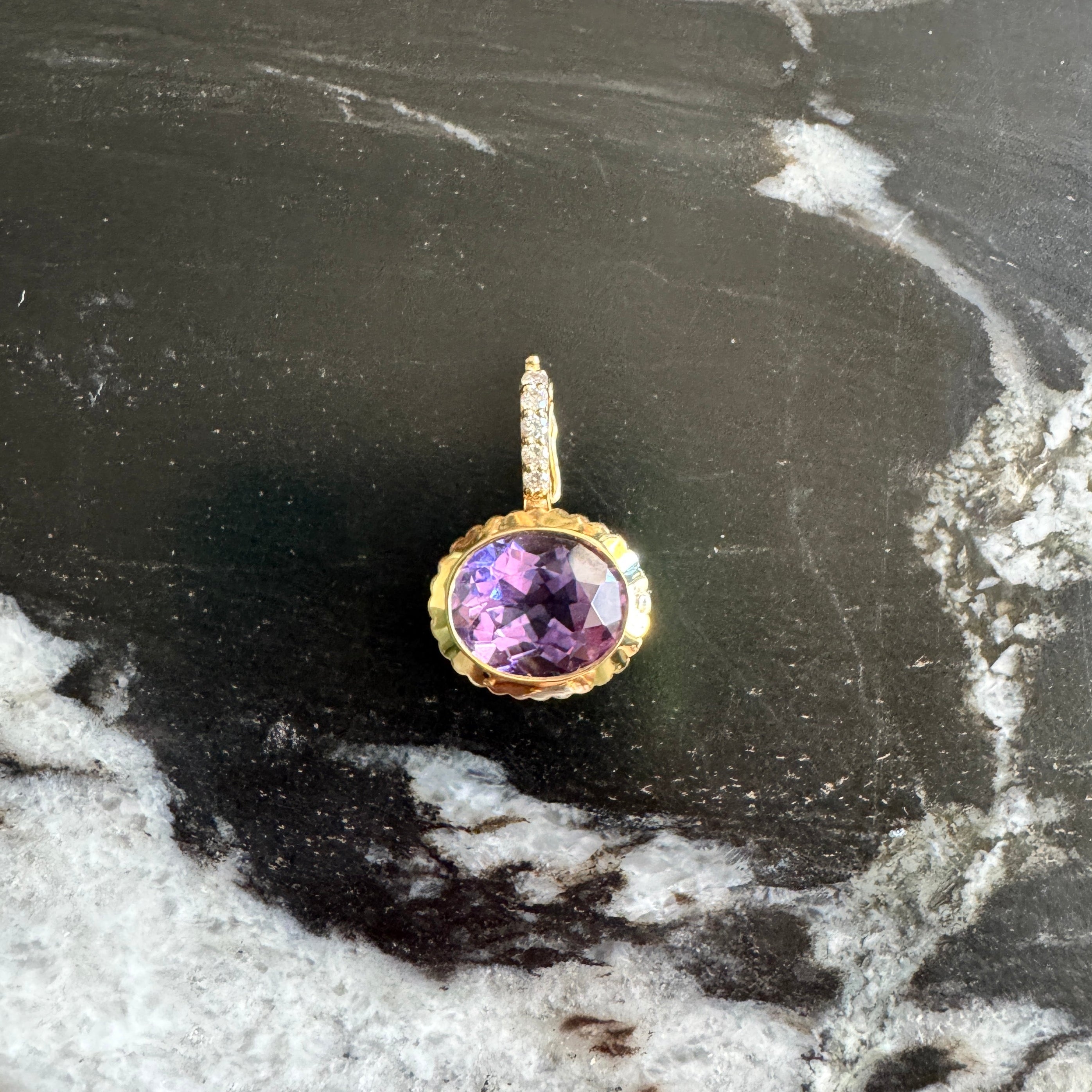 A gold charm with a round purple gemstone, displayed on a black and white marbled surface.