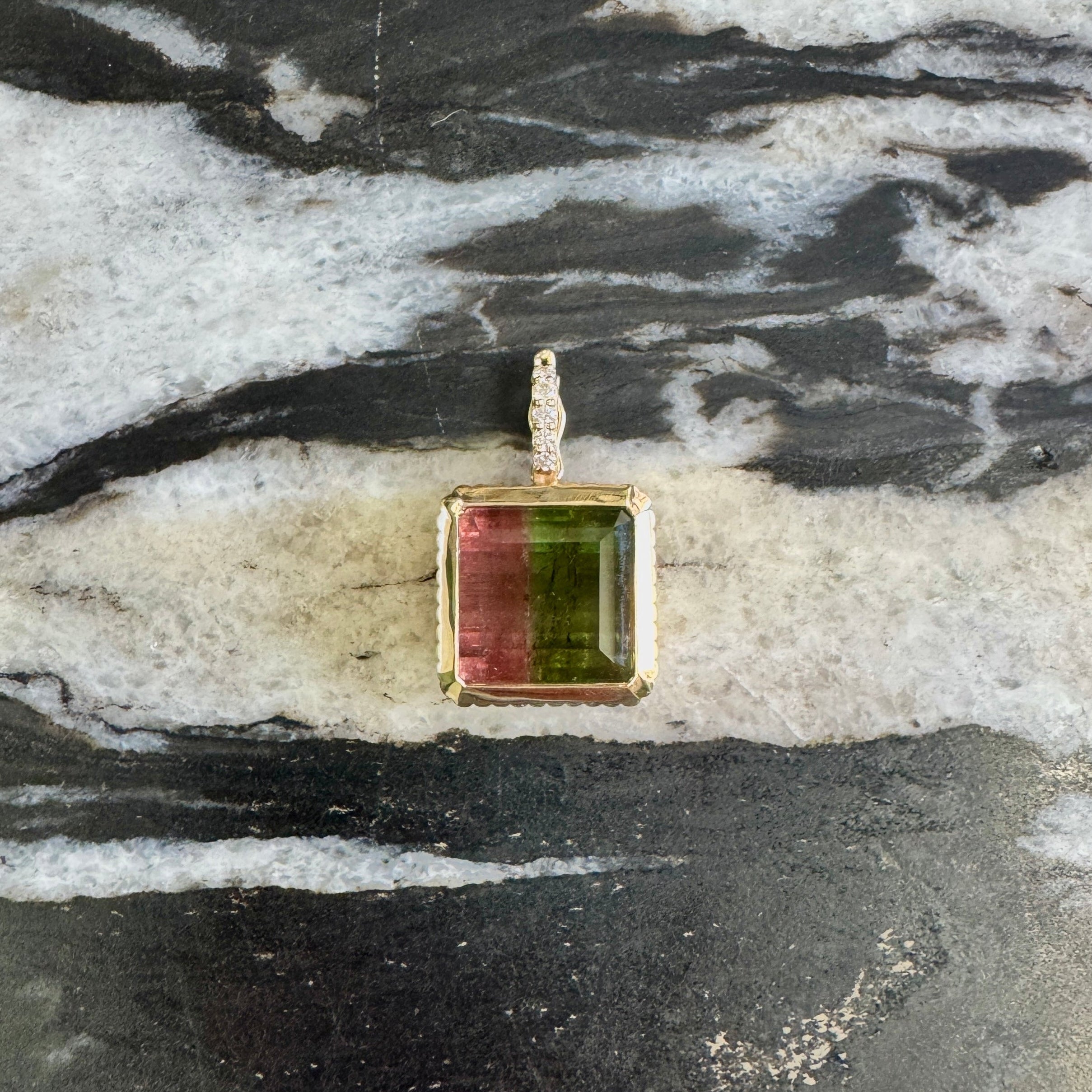 A square pendant with a gold-toned bezel setting featuring a green and pink banded stone, displayed on a black and white marbled surface.