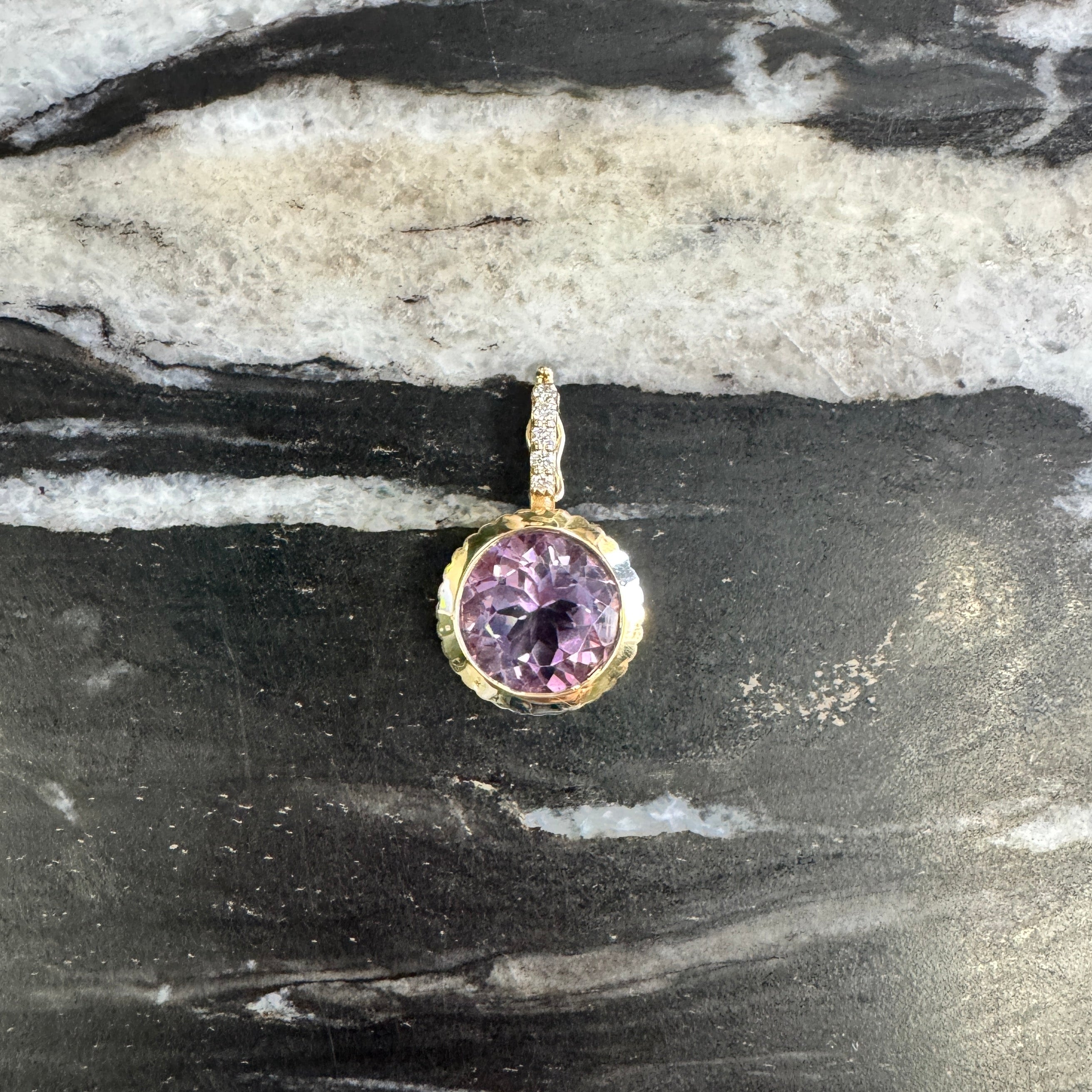 A small gold pendant with a round purple gemstone, resting on a black and white marbled surface.