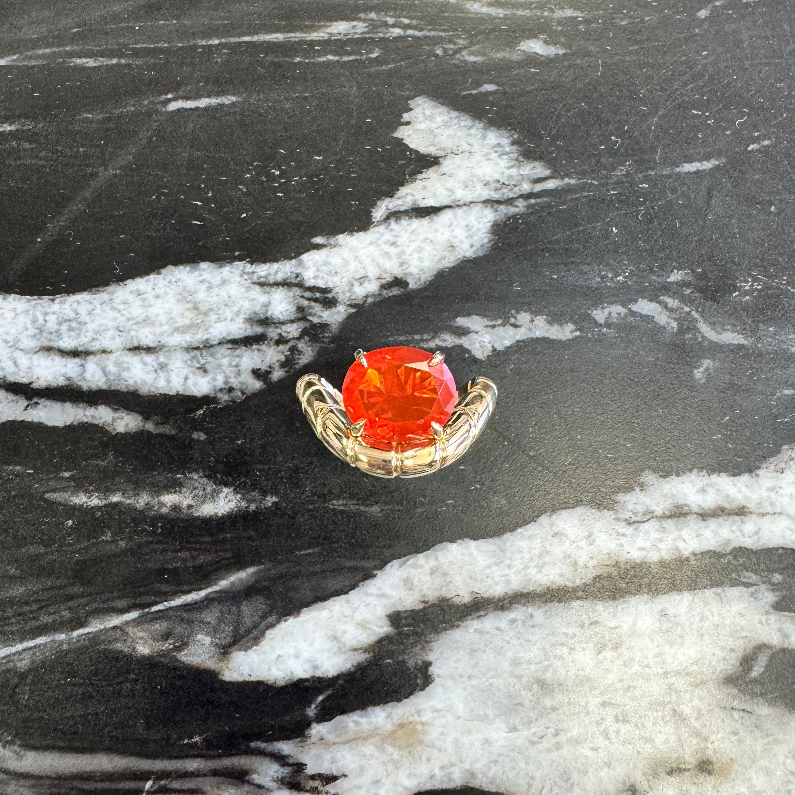 A small gold  ring with a round orange-red gemstone, displayed on a black and white marble surface.