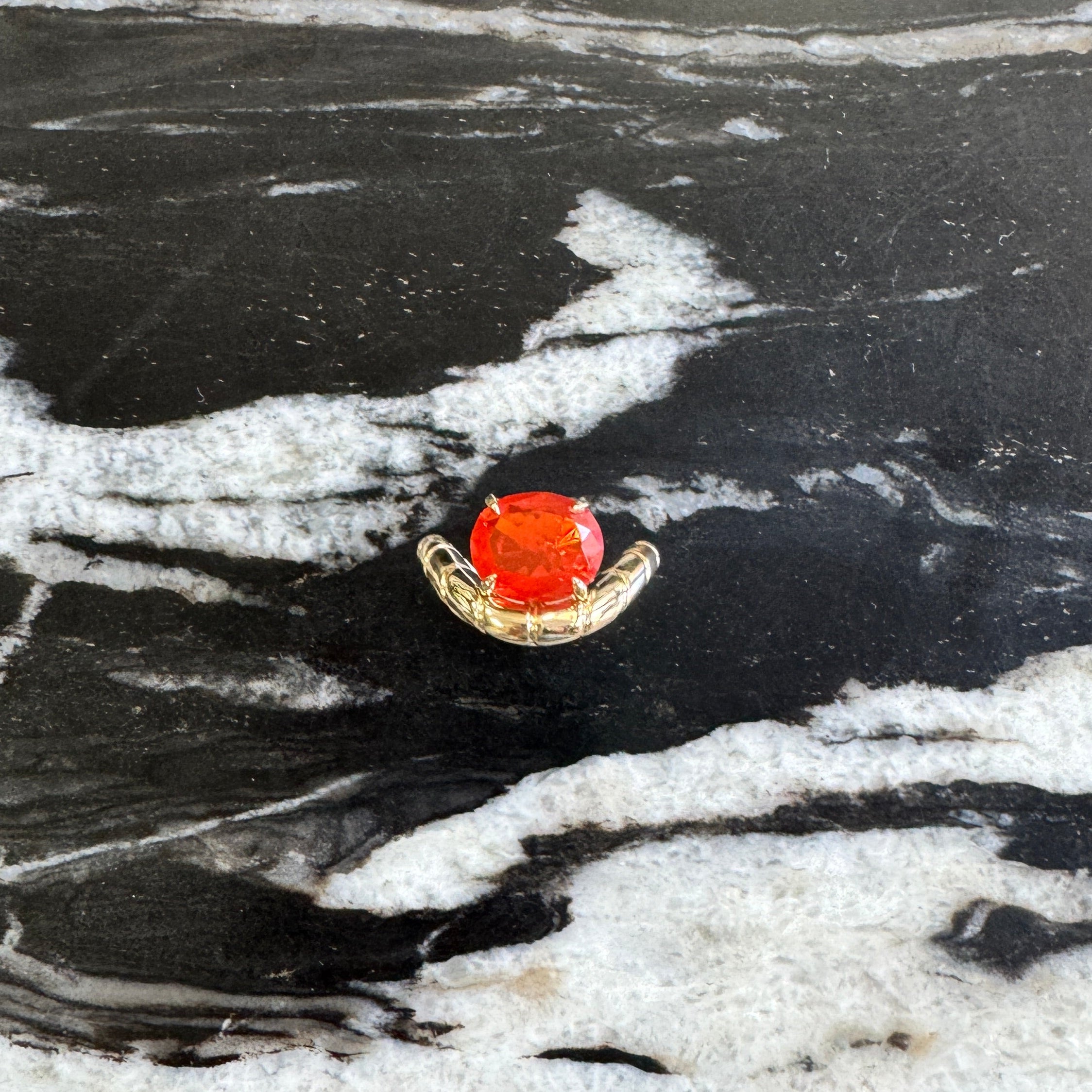 A small gold ringwith a round orange-red gemstone, displayed on a black and white marbled surface.