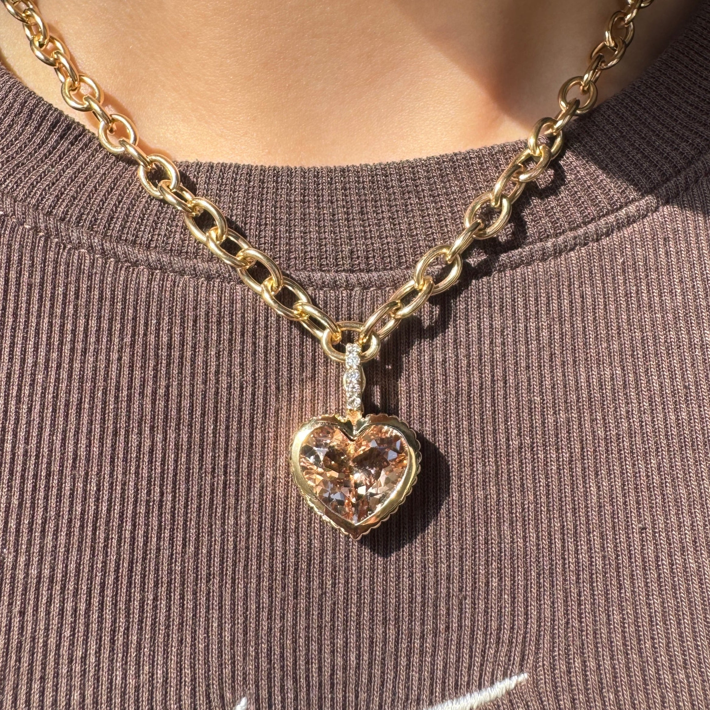 Person wearing a gold pendant with a peachy heart-shaped morganite and a diamond bail against a brown top 