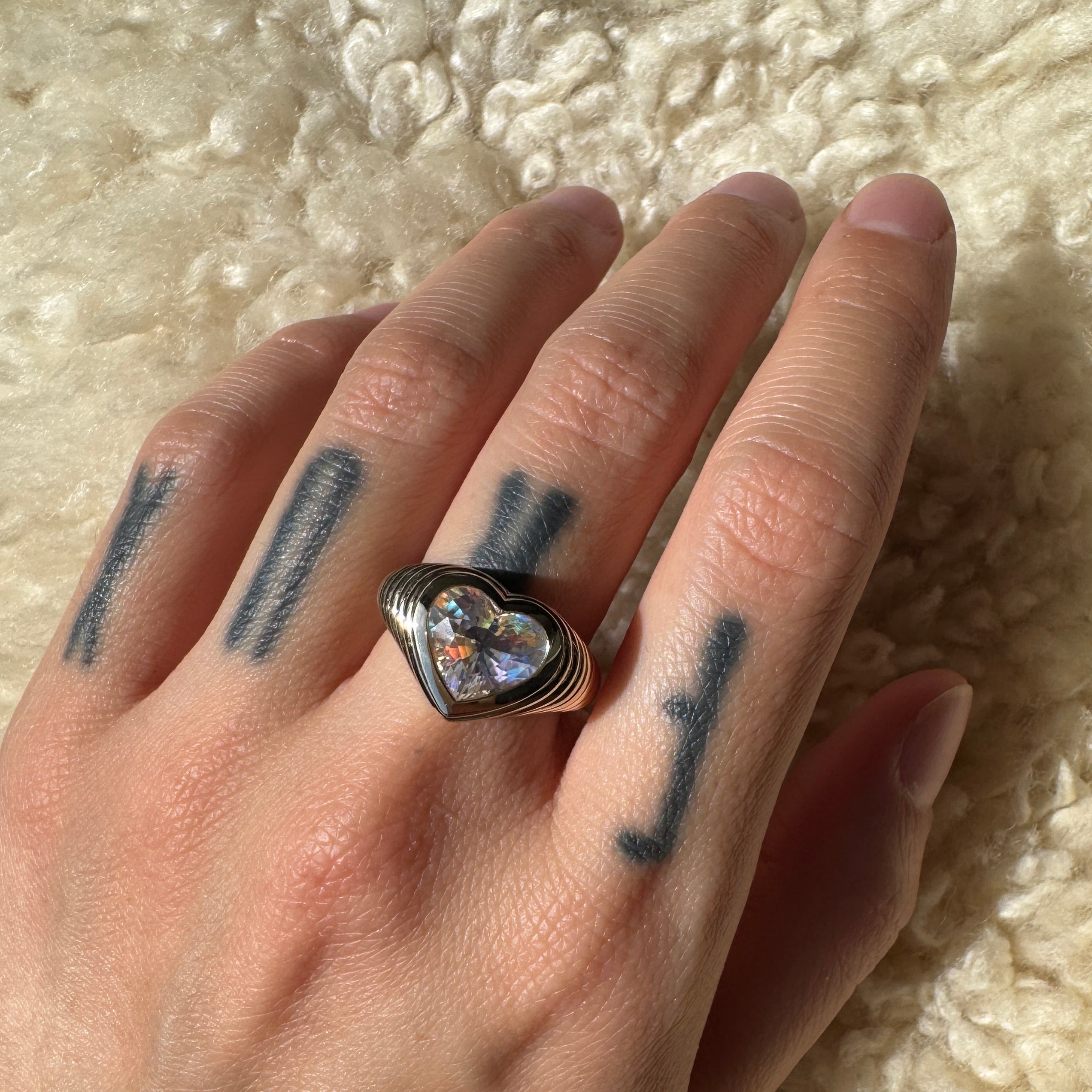 Hand wearing a gold ring with heart-shaped rainbow moonstone against a beige background 