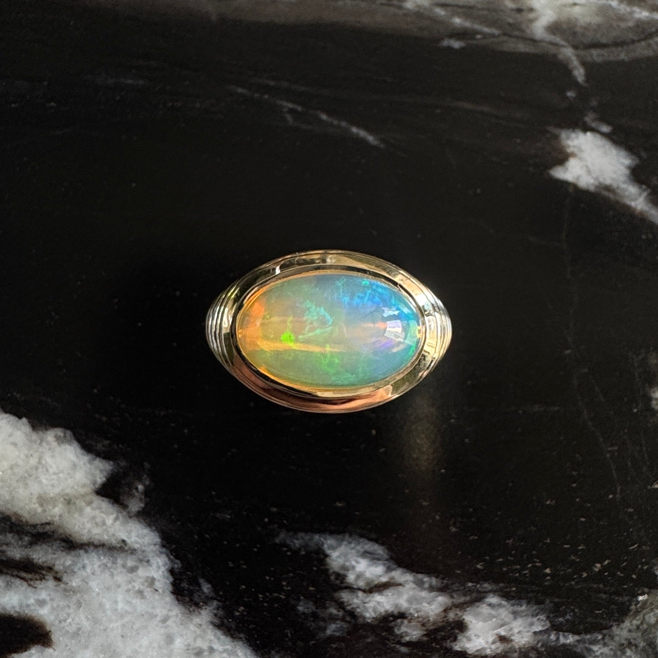Gold ring with an oval cabochon rainbow opal against a black and white marble background