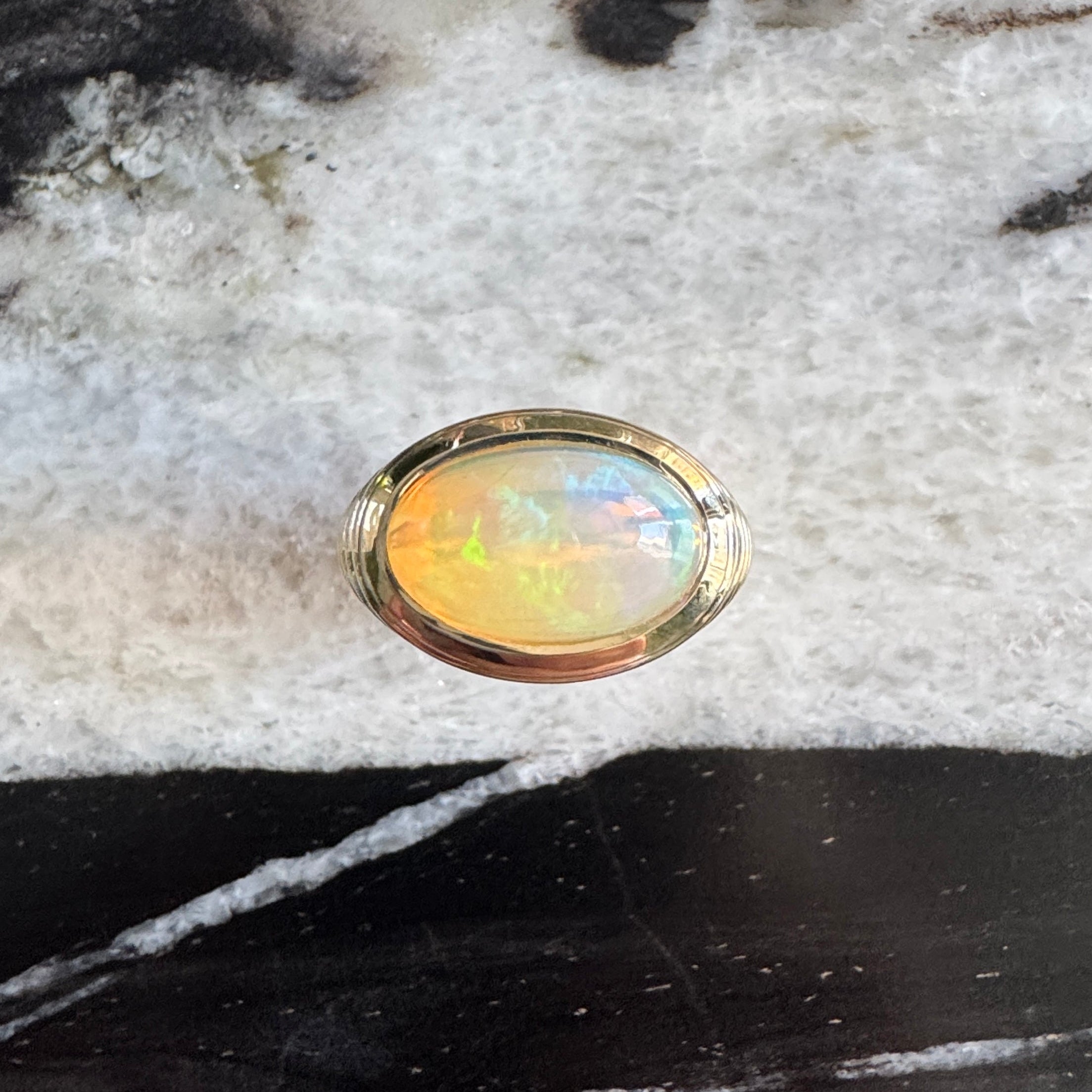 Gold ring with an oval cabochon rainbow opal against a black and white marble background