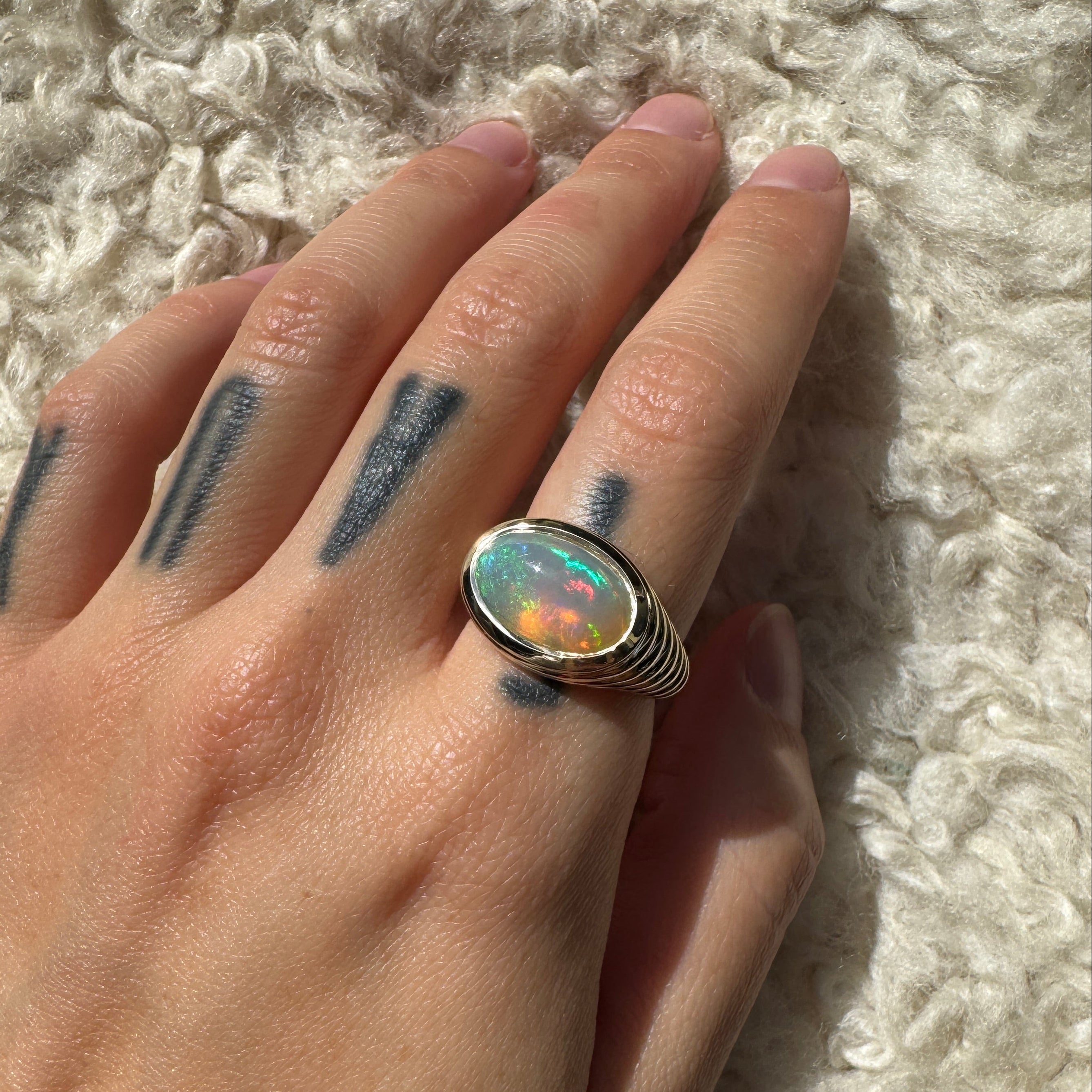 Hand wearing gold ring with oval cabochon rainbow opal against a beige background