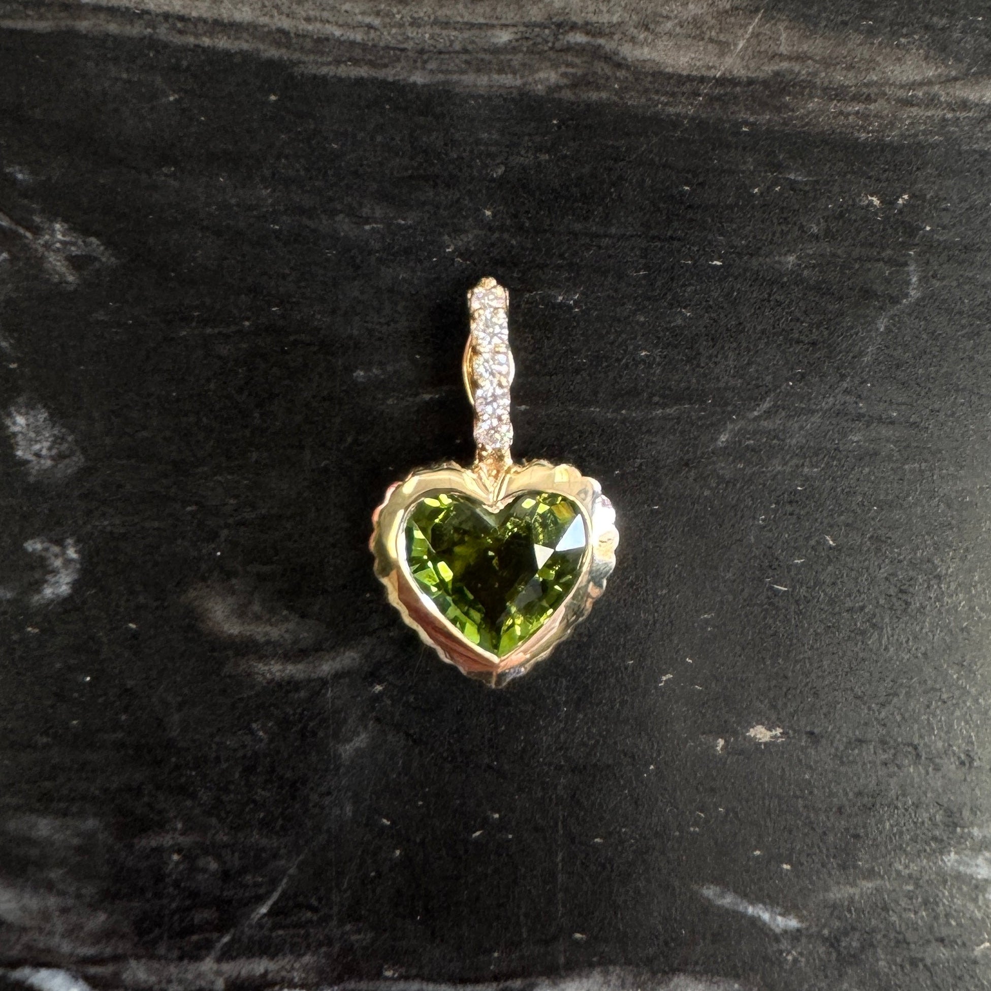 Gold pendant with a heart-shaped green tourmaline and a diamond bail against a black an white marble background 