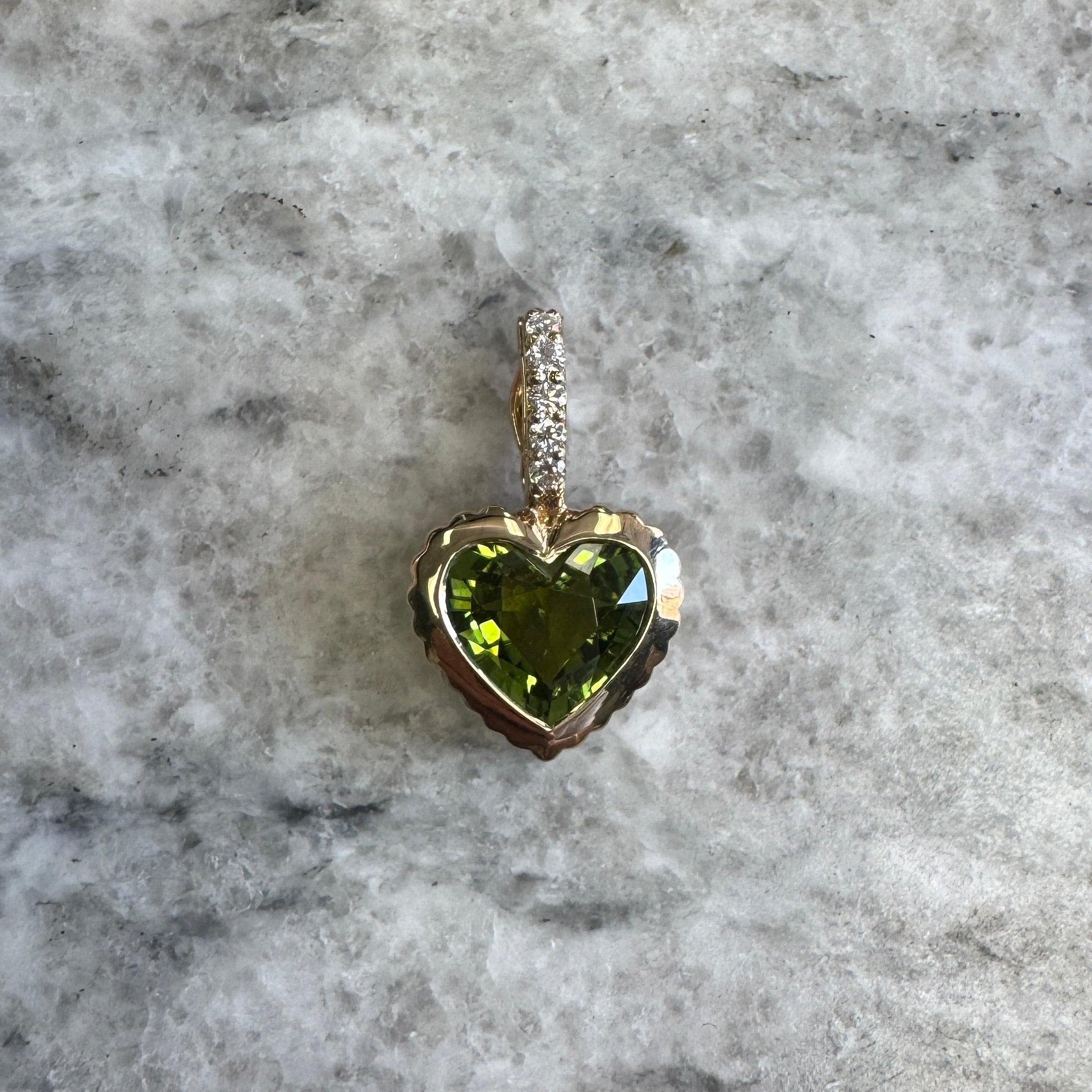 Gold pendant with a heart-shaped green tourmaline and a diamond bail against a black an white marble background 