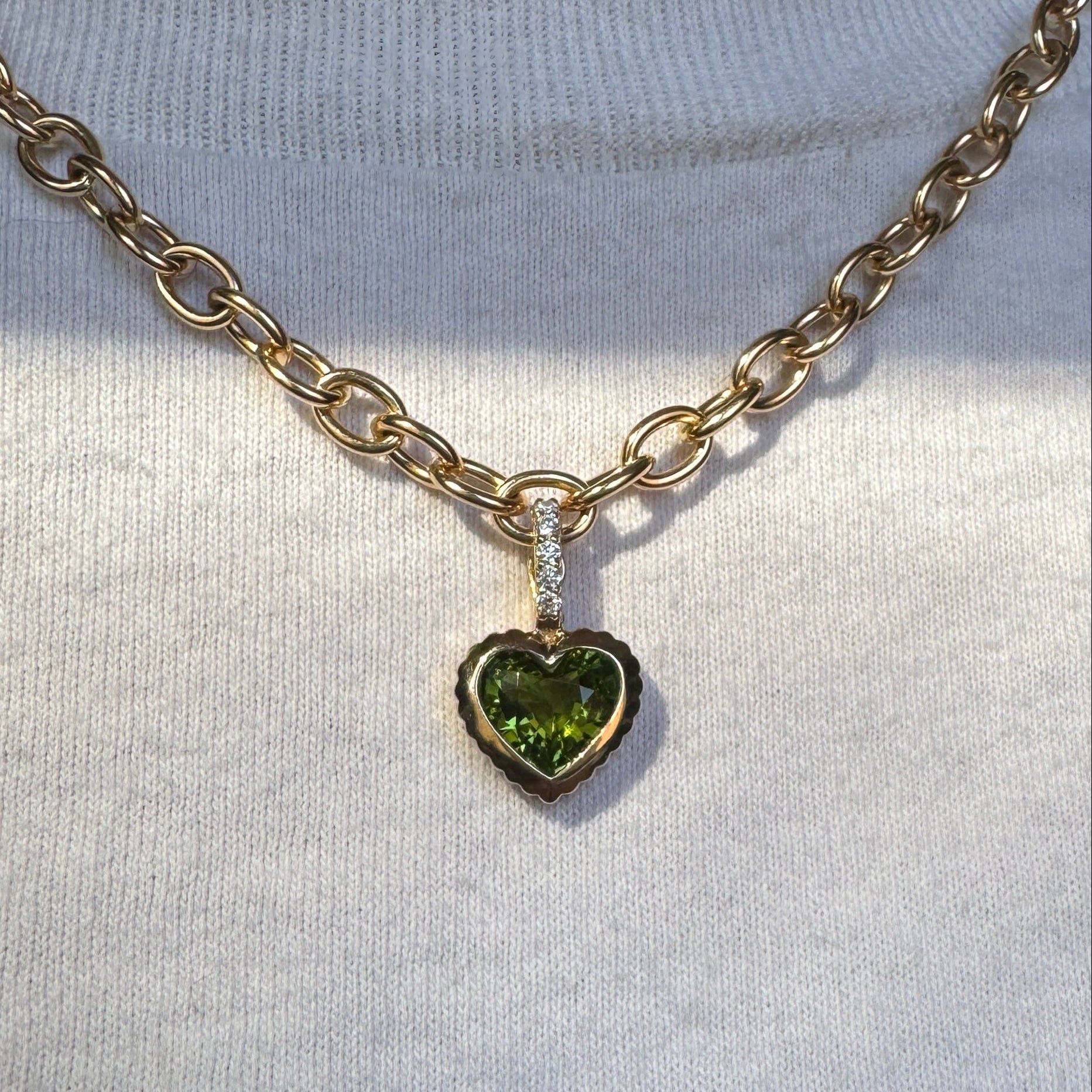 Person wearing a gold pendant with a heart-shaped green tourmaline and a diamond bail on a gold chain against a white top