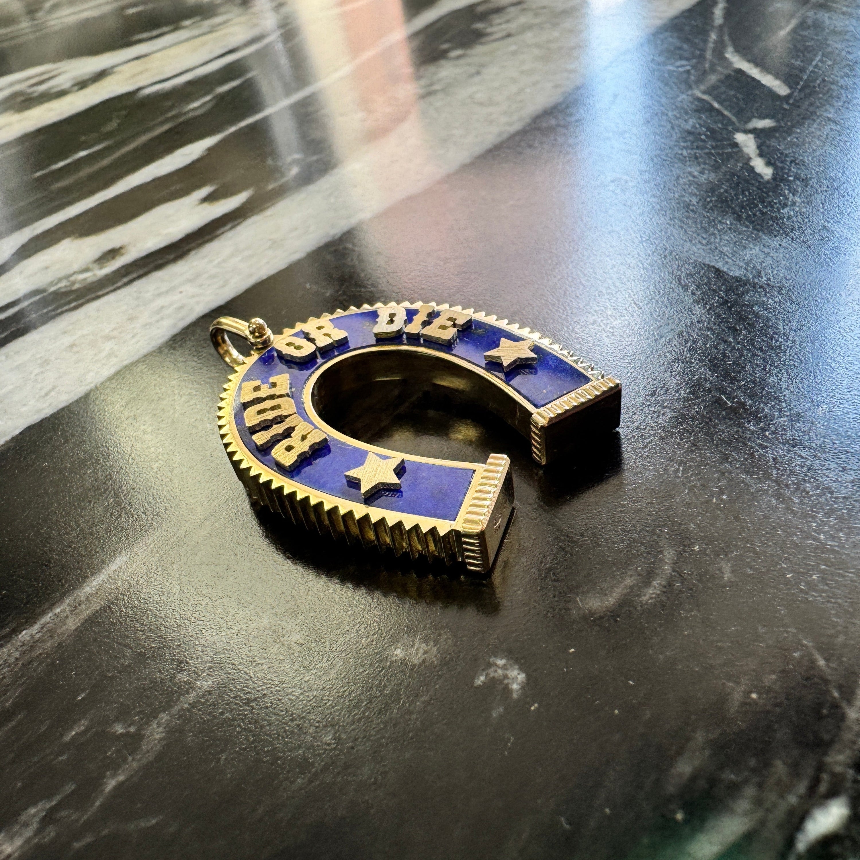 A gold horseshoe-shaped pendant with blue lapis and raised gold lettering spelling 'Ride or Die' with star details, resting on a dark marble surface.