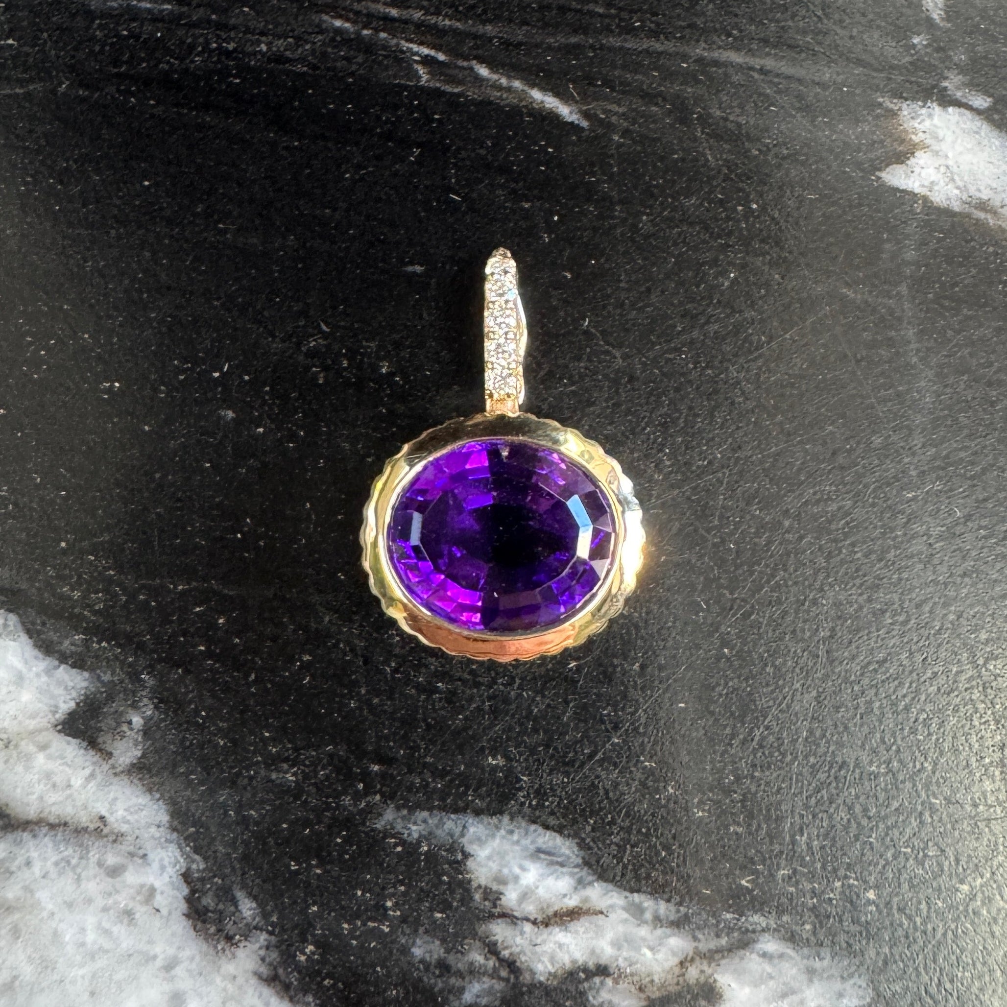 Gold pendant with an oval purple amethyst and a diamond bail against a black and white marble background