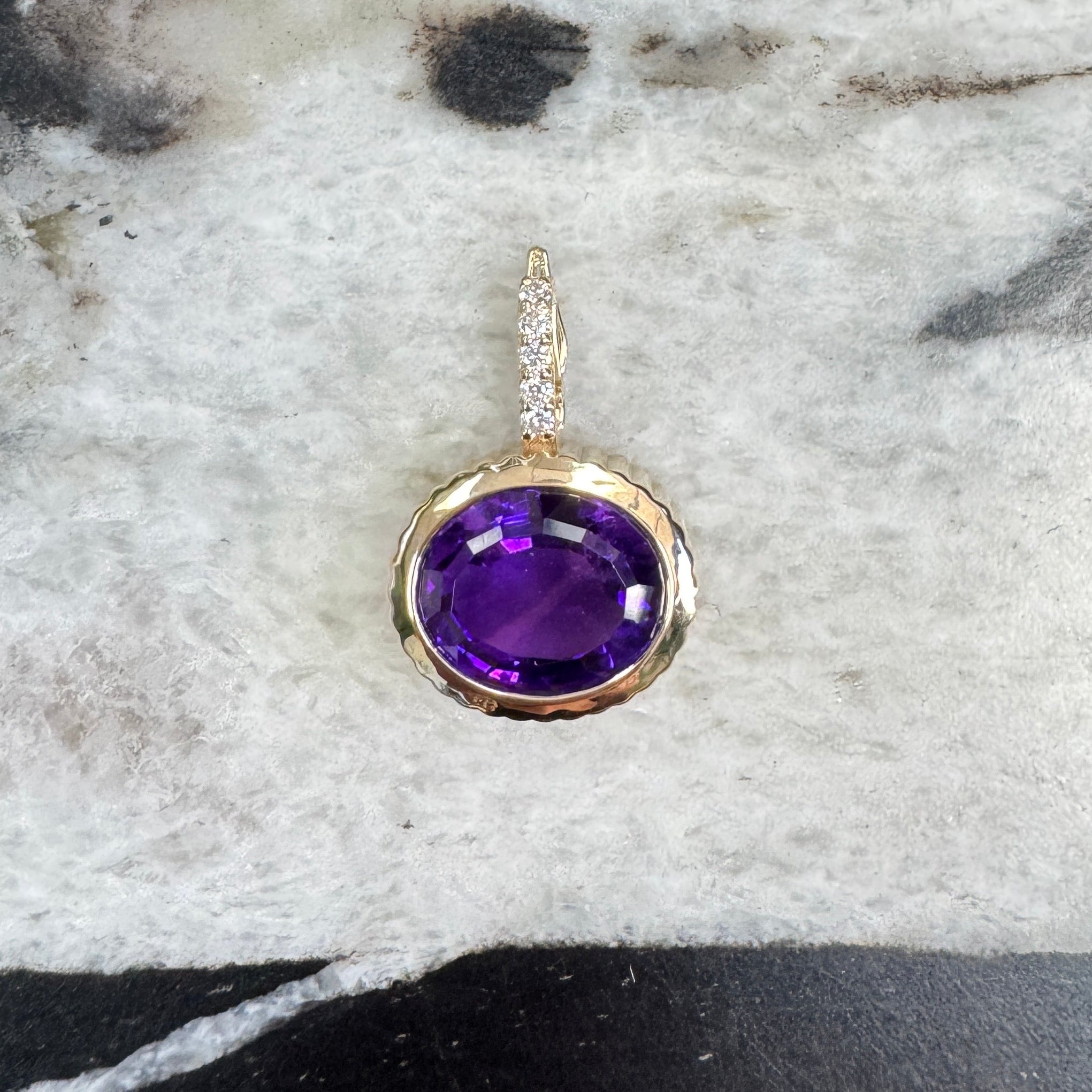 Gold pendant with an oval purple amethyst and a diamond bail against a black and white marble background