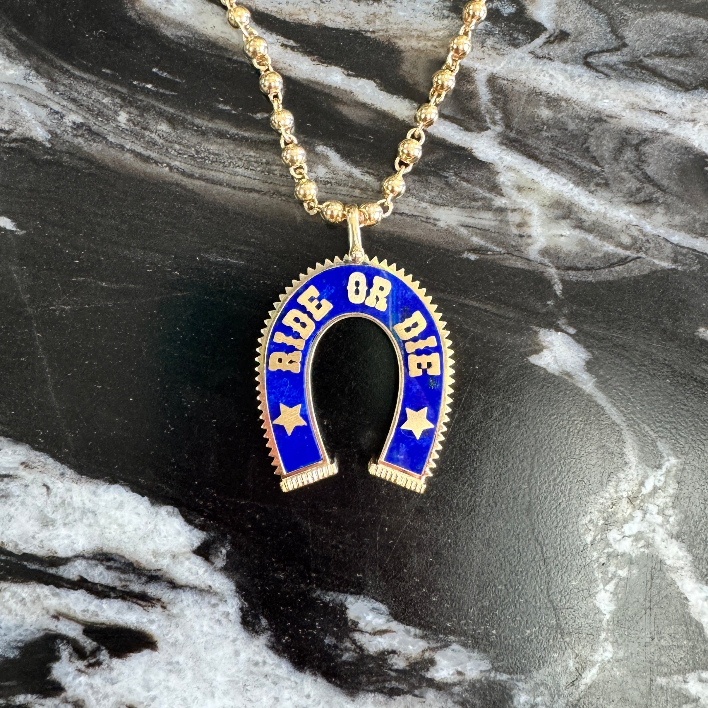 A gold-tone chain necklace with a horseshoe-shaped pendant featuring blue lapis and the words 'RIDE OR DIE' in gold, displayed on a black and white marble surface.