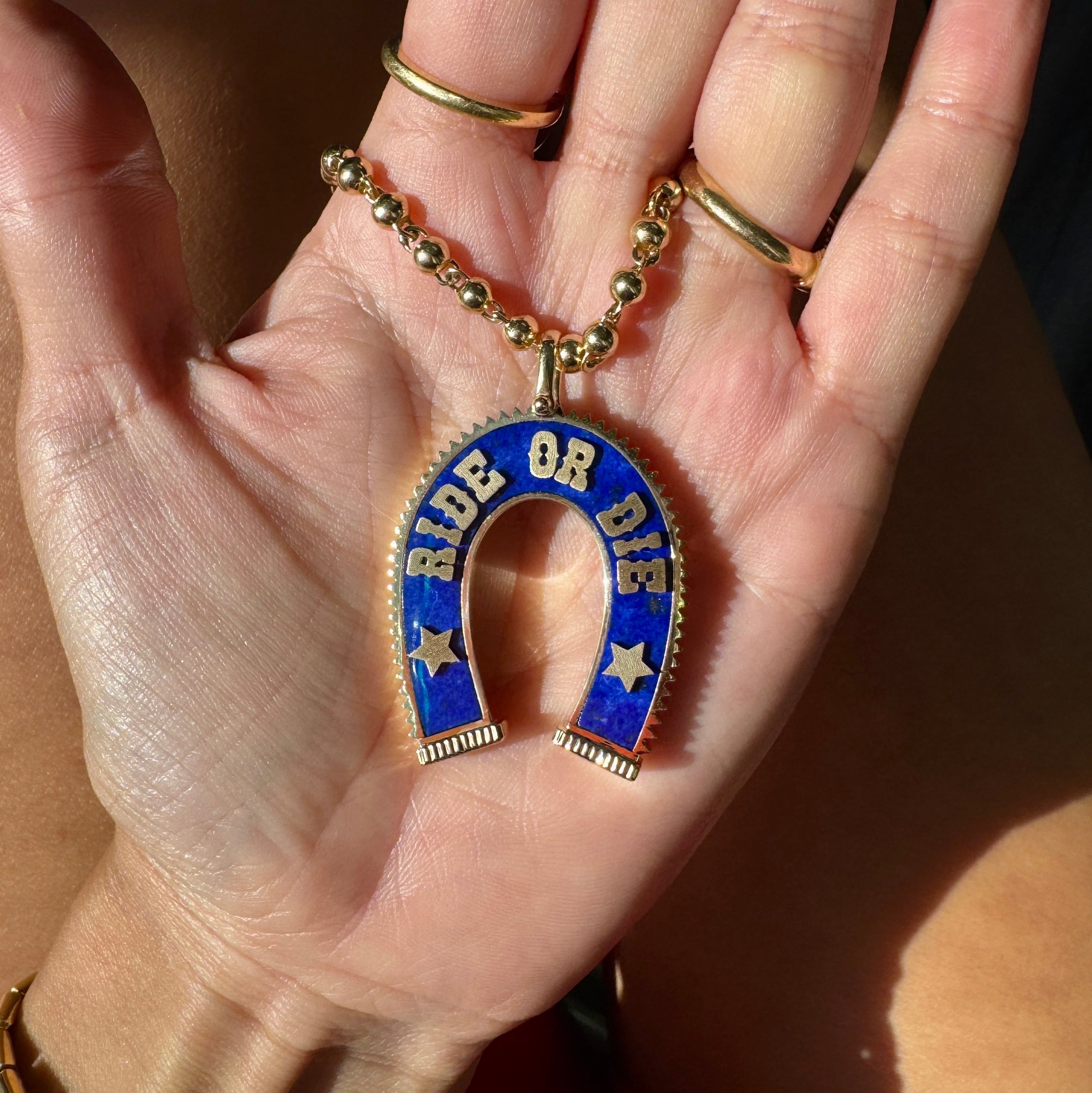 A gold horseshoe pendant necklace with blue lapis and white lettering that reads 'RIDE OR DIE, held in a person's open hand. The necklace is on a gold ball chain, and the person is wearing gold rings and a gold watch.