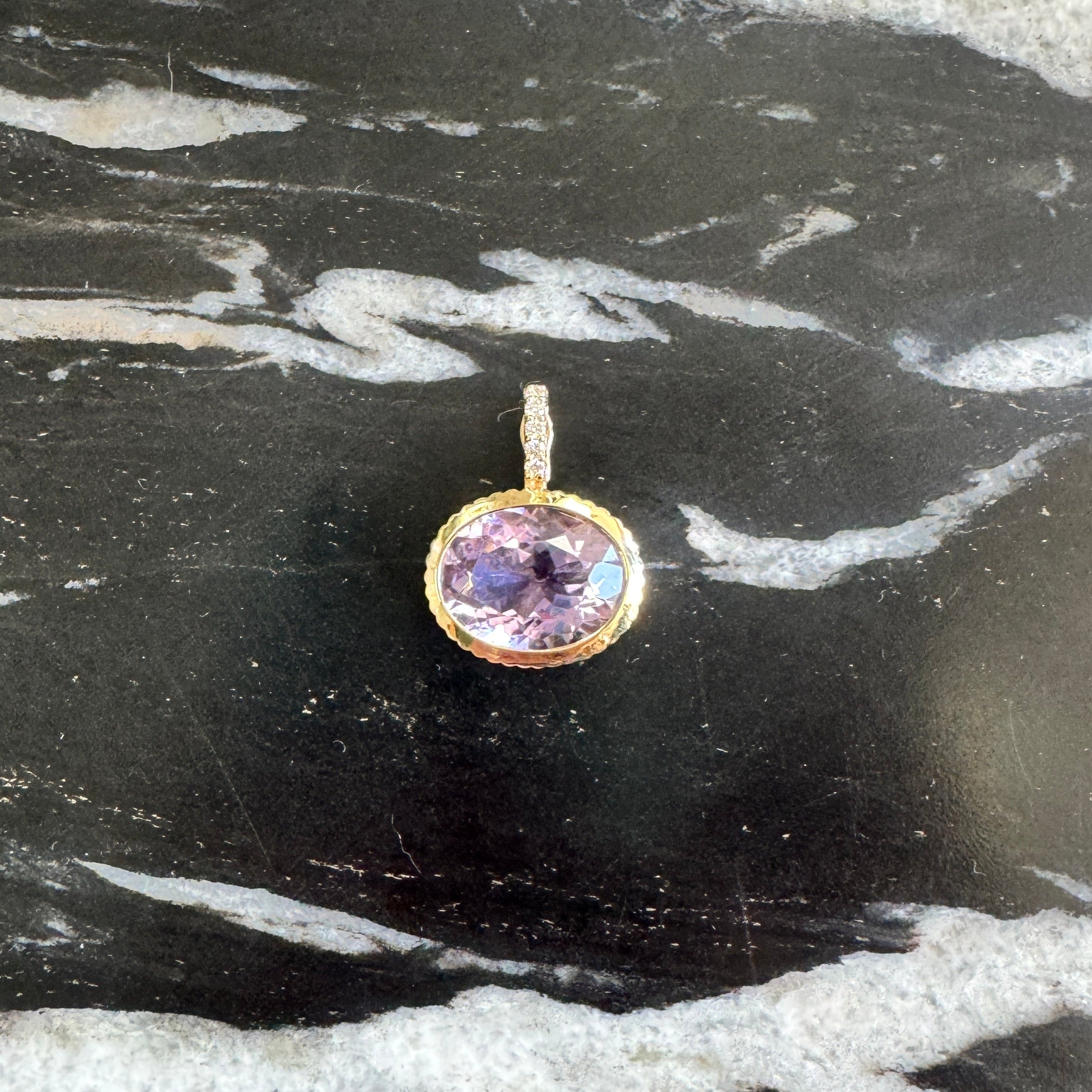 A small oval pendant with a purple gemstone in a gold bezel setting, placed on a black and white marbled surface.