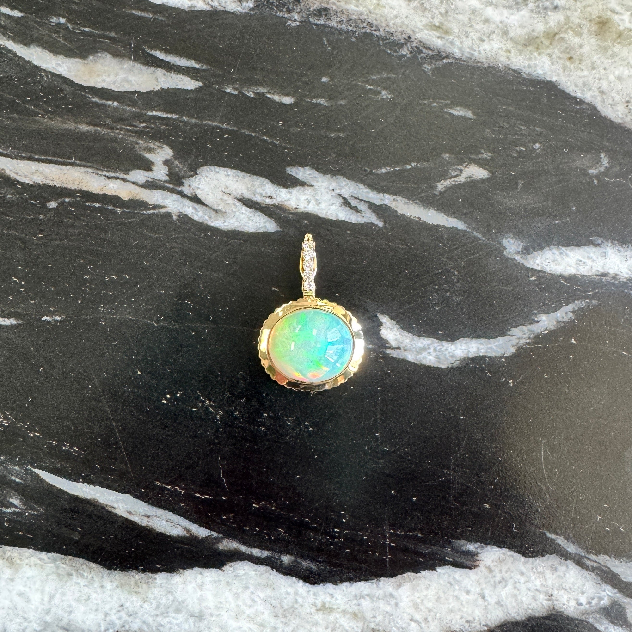 A gold pendant with an oval green-blue opal stone, resting on a black and white marbled surface.