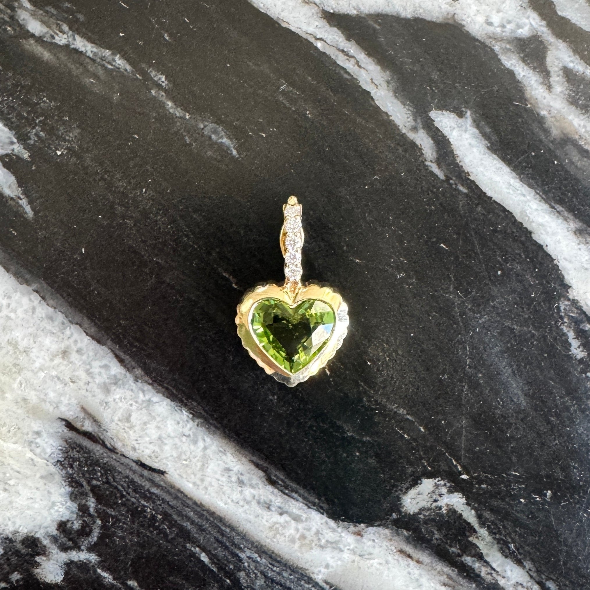 A small heart-shaped pendant with a green gemstone and gold-tone setting, photographed on a black and white marbled surface.