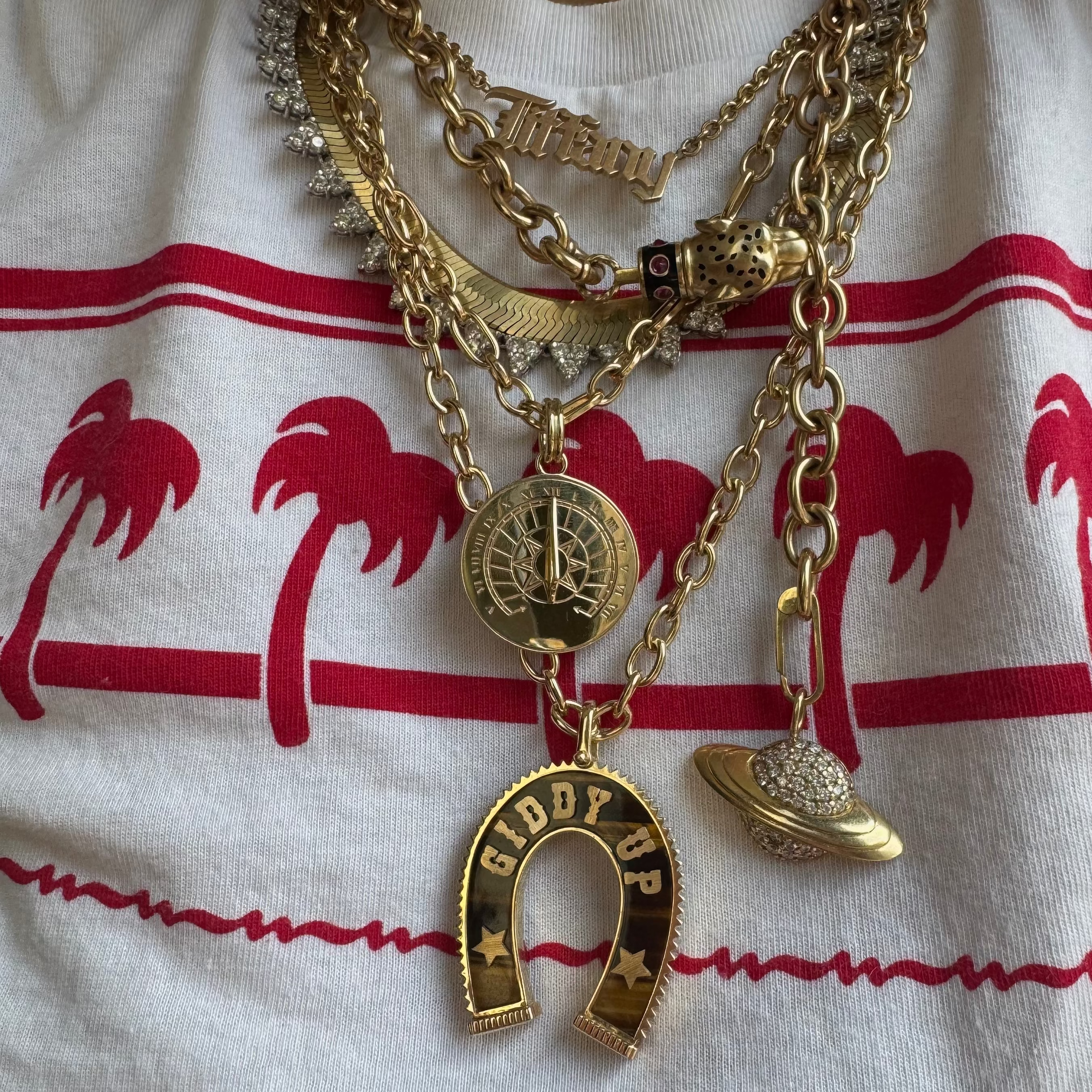 A close-up of a person wearing a layered gold necklace set over a white t-shirt with red palm tree graphics. The necklace features multiple chains, a 'Tiffany' nameplate,