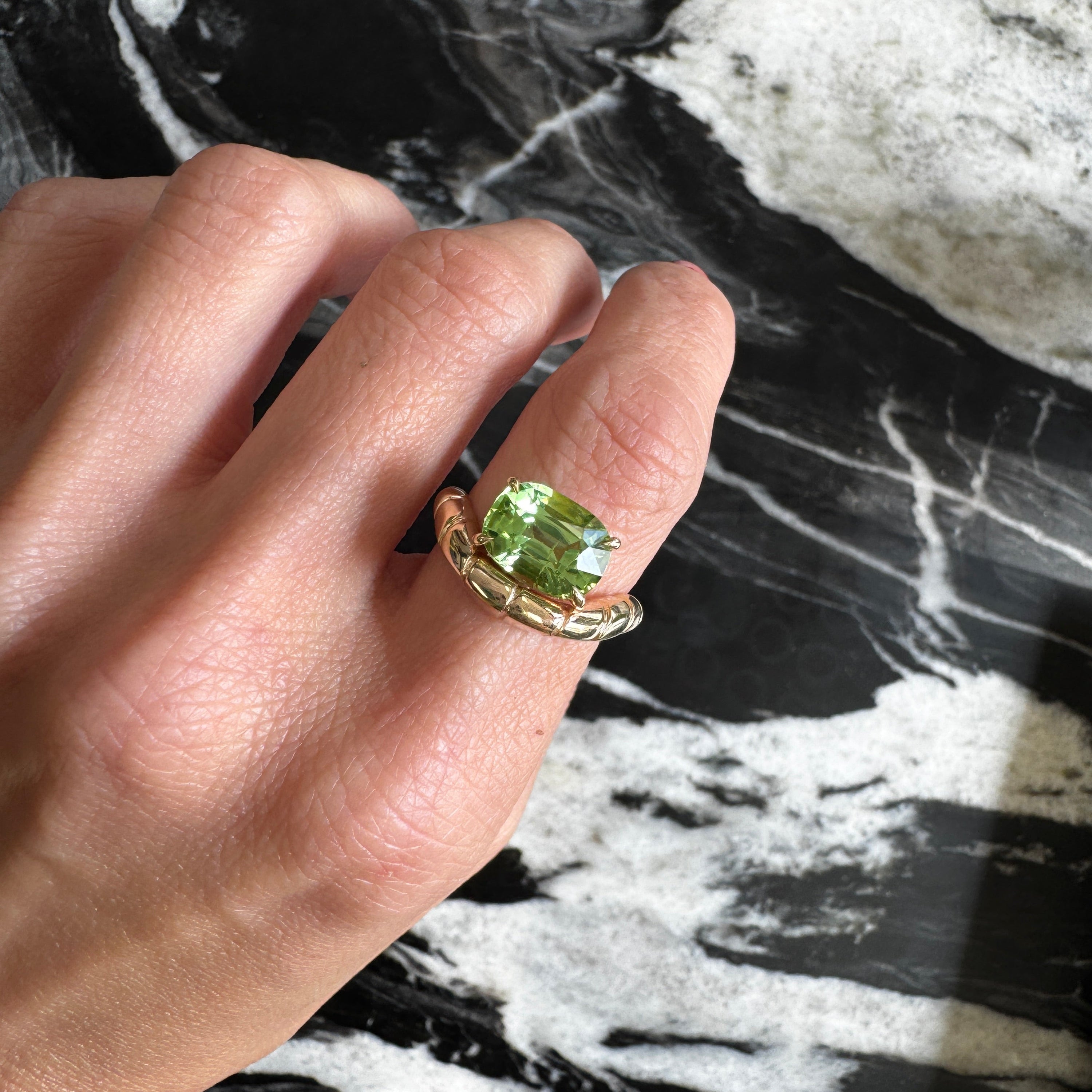 Hand wearing a gold ring with a green gemstone against a marble background